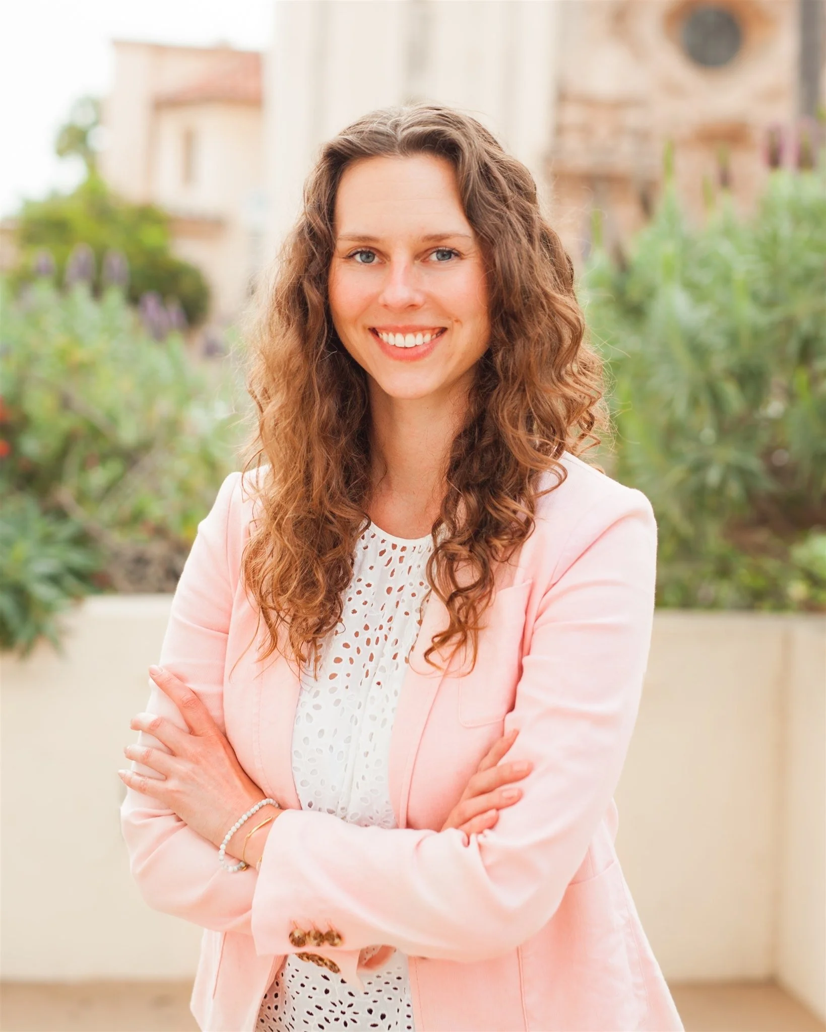Woman smiling in a pink blazer, standing in an outdoor setting with greenery in the background.