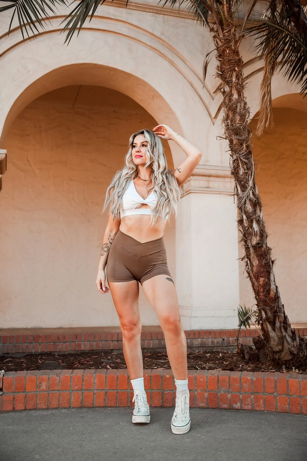 Woman in workout clothing standing under an arch next to a palm tree, smiling and looking upwards.