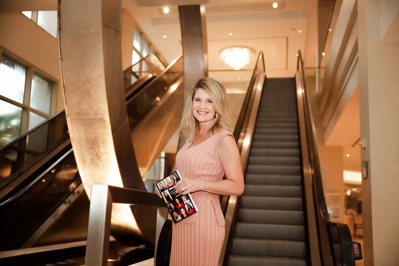 Woman in pink dress holding a book, standing near an escalator in a modern indoor setting.