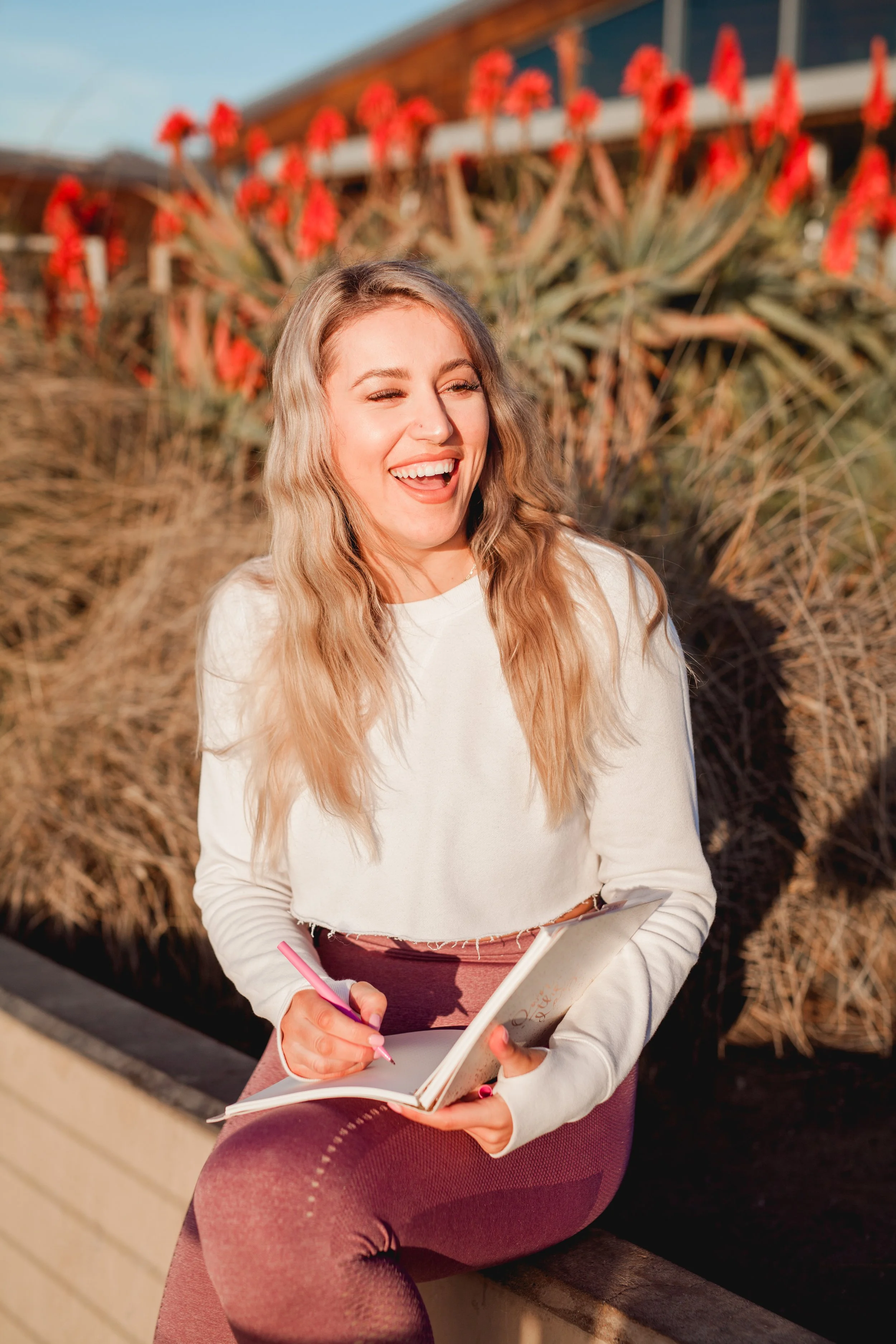 Woman smiling and writing in a notebook outdoors with red aloe plants in the background.