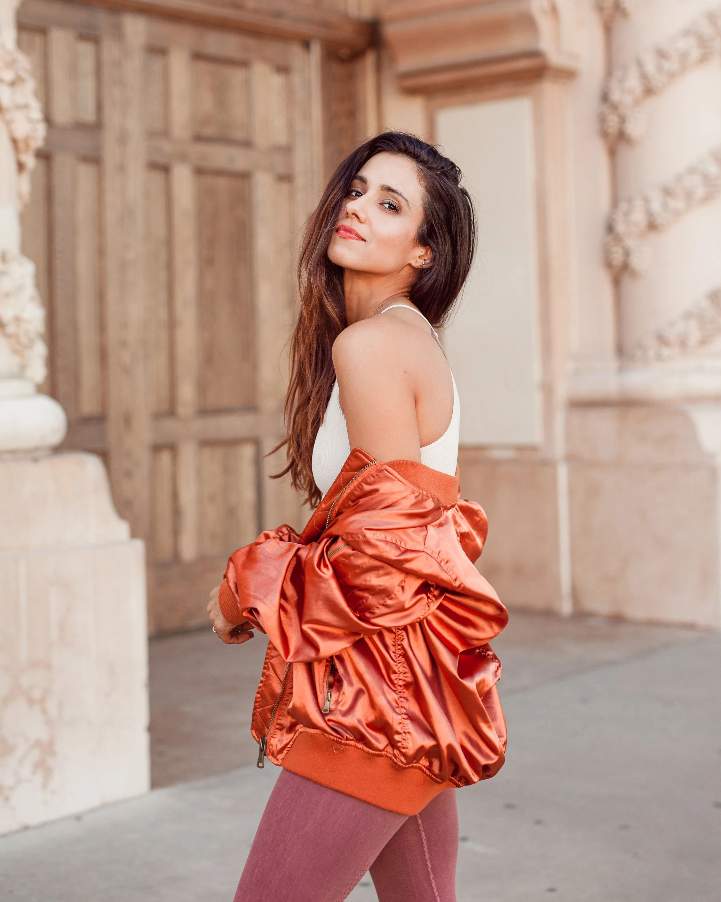 Woman posing with a relaxed smile, wearing an orange jacket over her shoulder, in front of an ornate doorway.