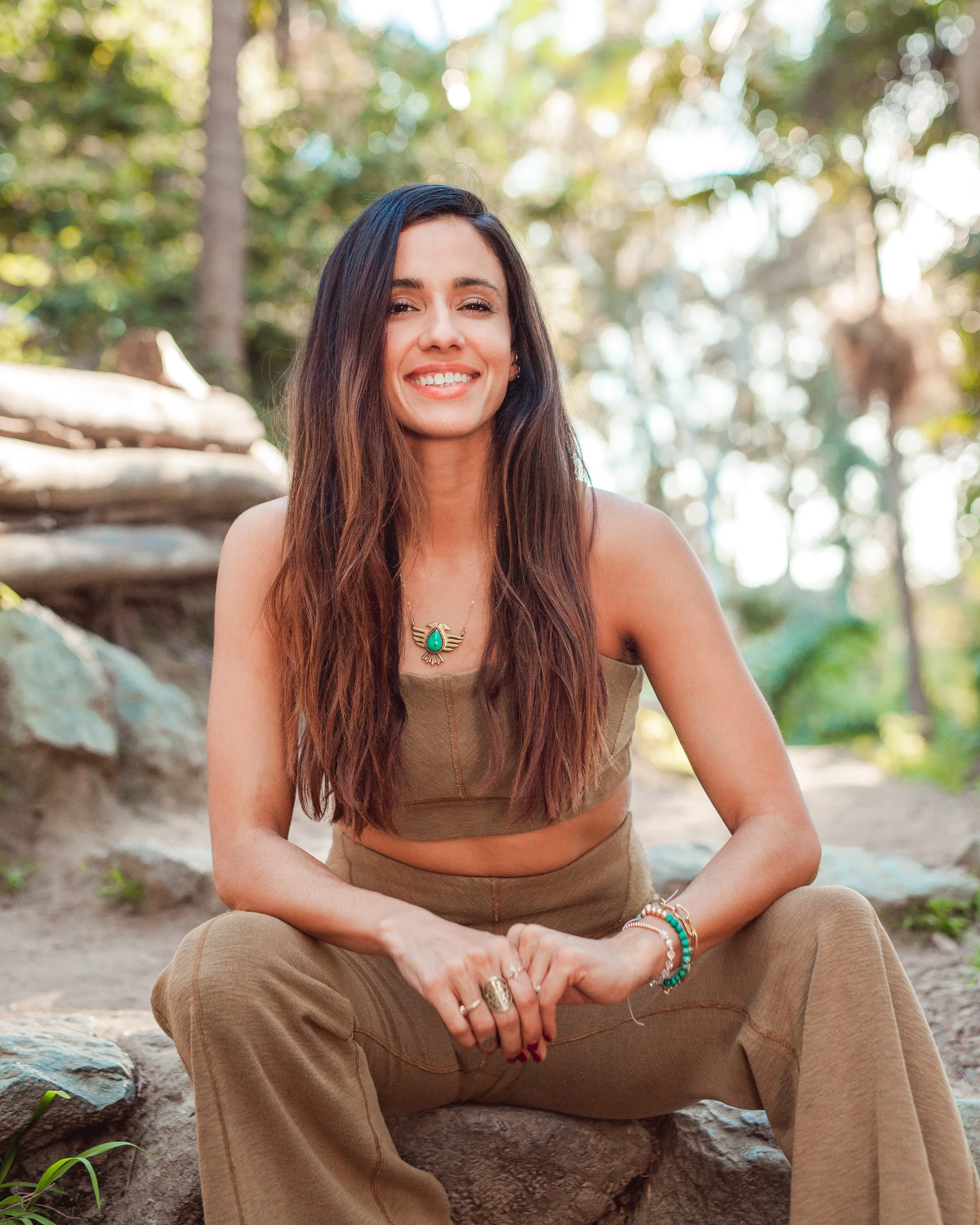 Woman seated outdoors wearing a brown crop top and pants, smiling with long hair, surrounded by greenery and trees.
