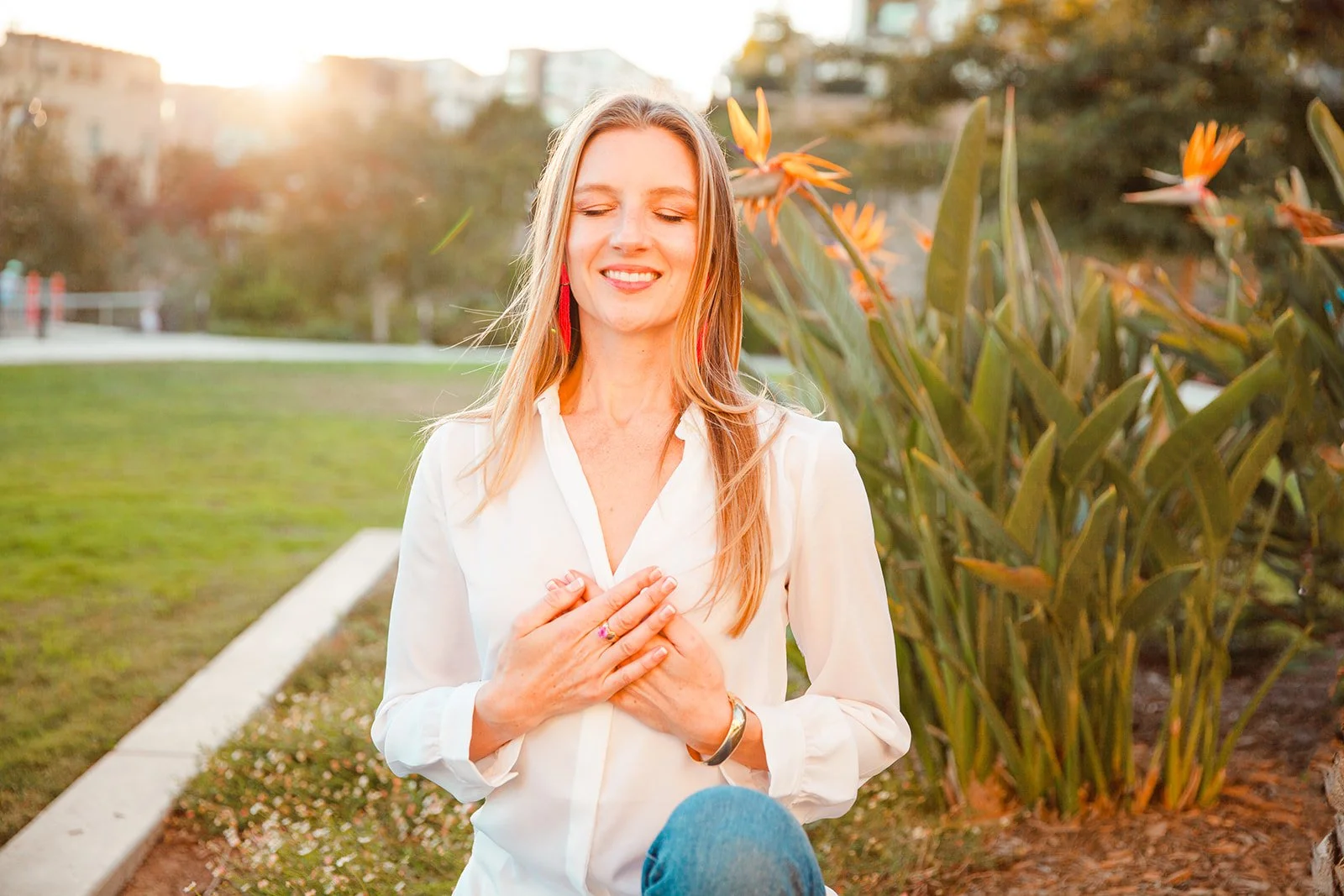 Woman meditating in a garden with Bird of Paradise flowers, wearing a white blouse and sitting in a peaceful outdoor setting, sunlight in background.