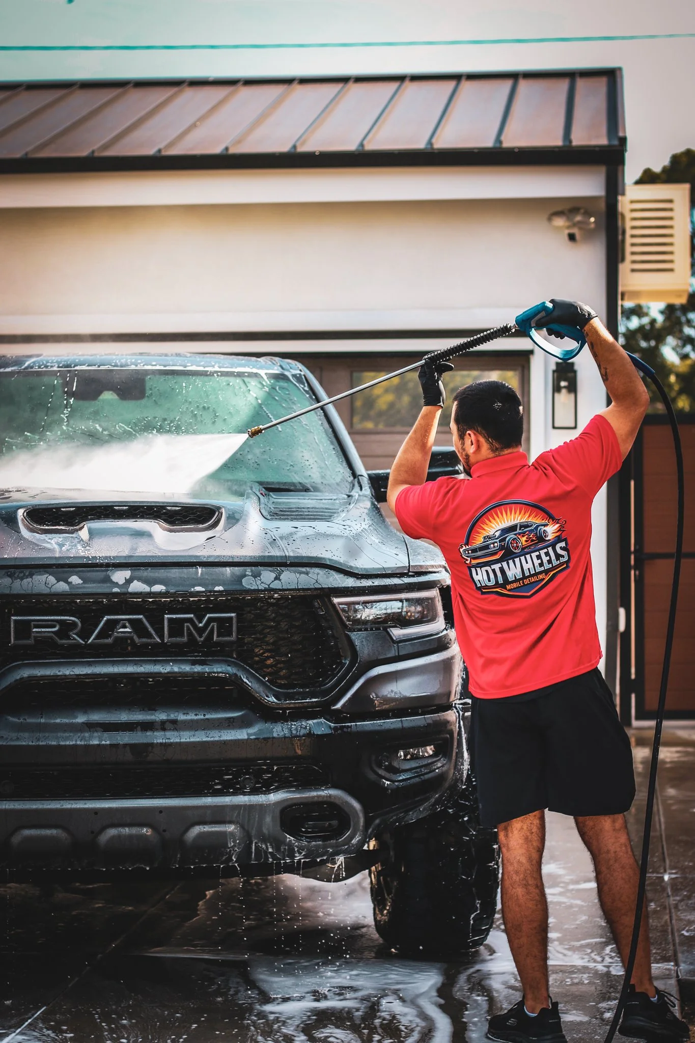 A man wearing a red shirt with a car wash logo is washing a black RAM truck with a pressure washer at a residential driveway.