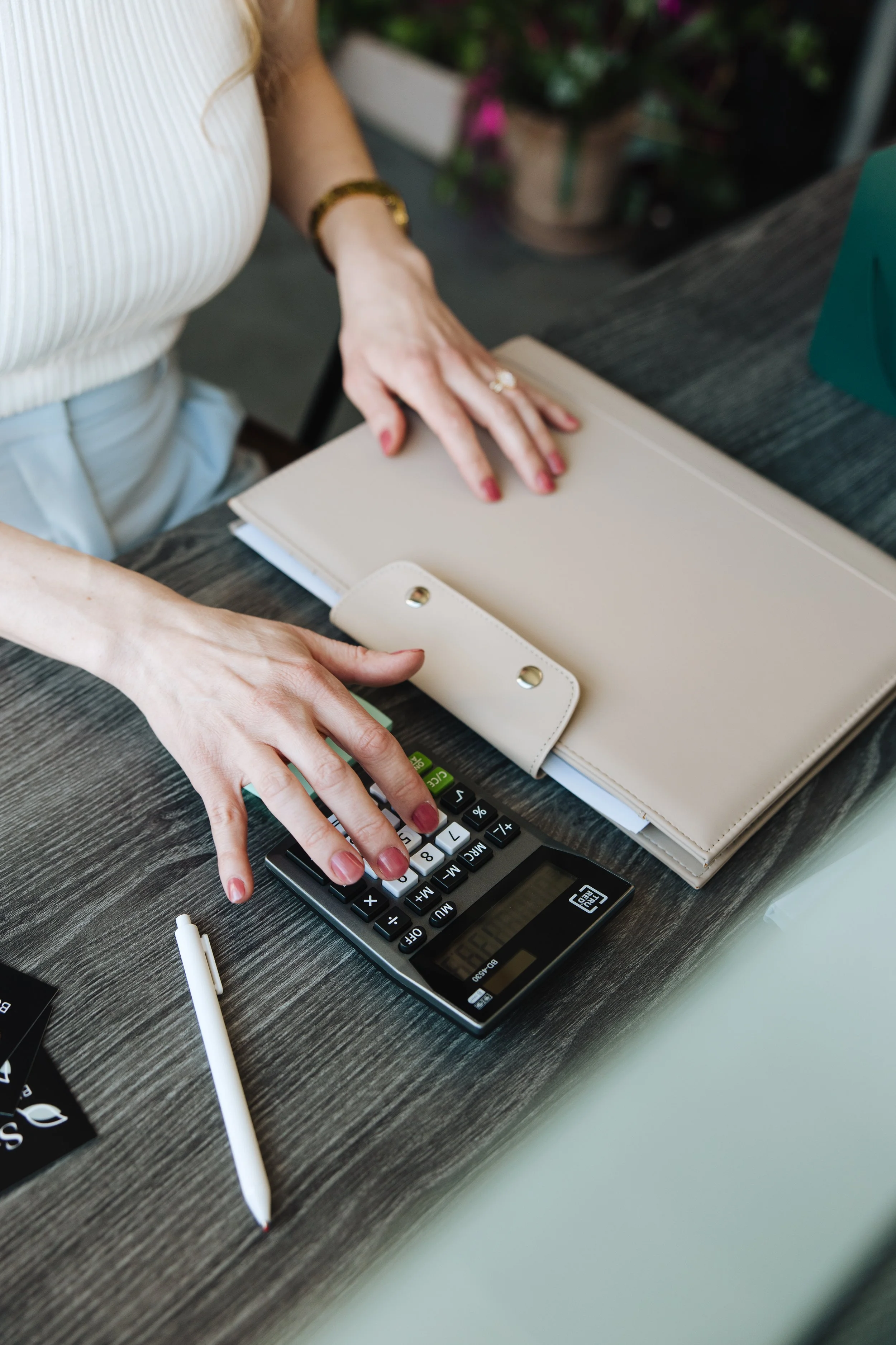 Person using a calculator on a desk with a beige planner, white pen, and some business cards.
