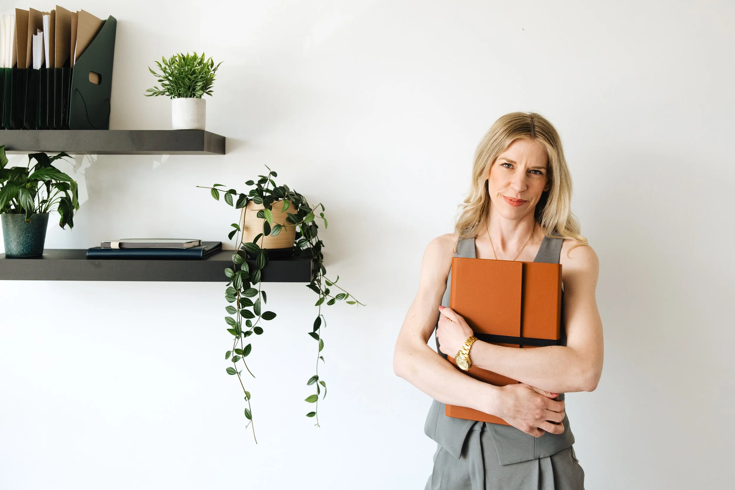 A woman with blonde hair in a gray sleeveless dress holding an orange notebook, standing in front of a white wall with green potted plants and shelves with books and folders.