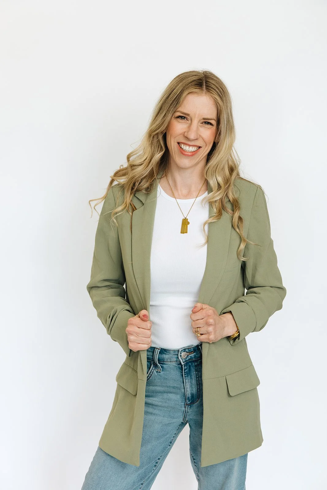 A smiling woman with long wavy blonde hair wearing a green blazer, white t-shirt, and blue jeans, standing against a white background.