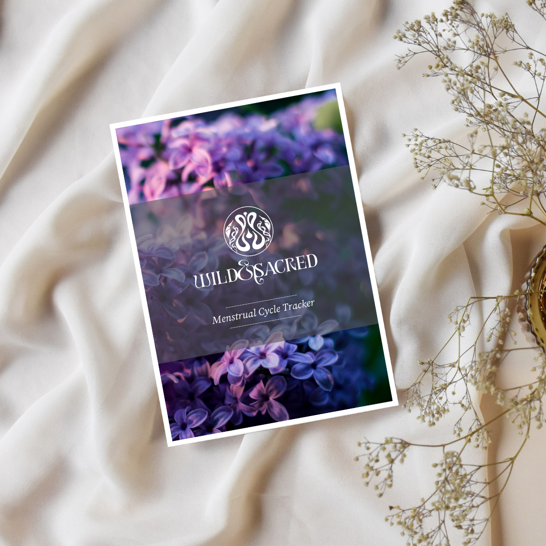 Cover of "Wild & Sacred: Menstrual Cycle Tracker" featuring purple flowers and decorative text on a white fabric backdrop, with dried flowers nearby.