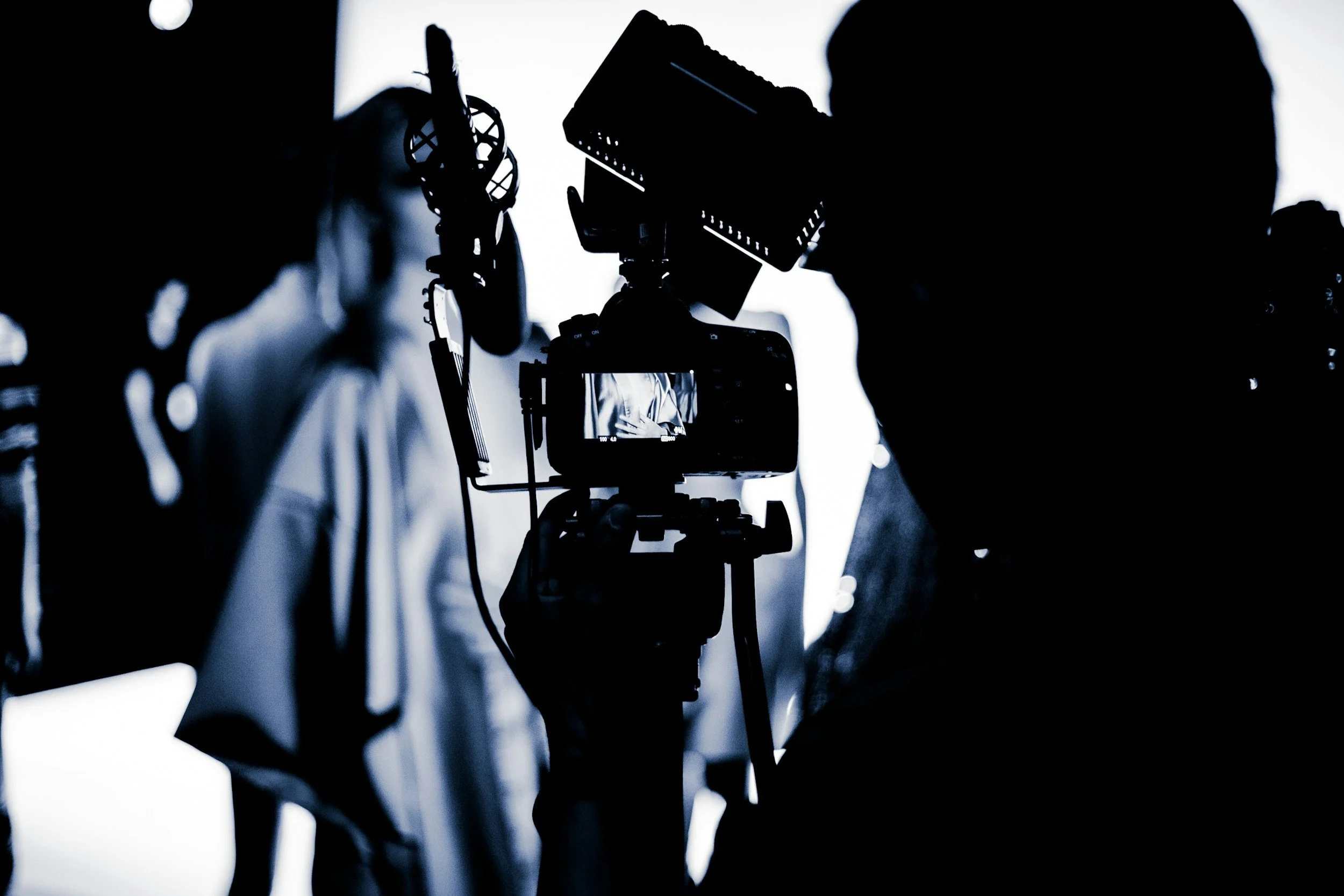 Silhouette of a person operating a video camera in a studio setting, with blurred background figures and professional lighting.