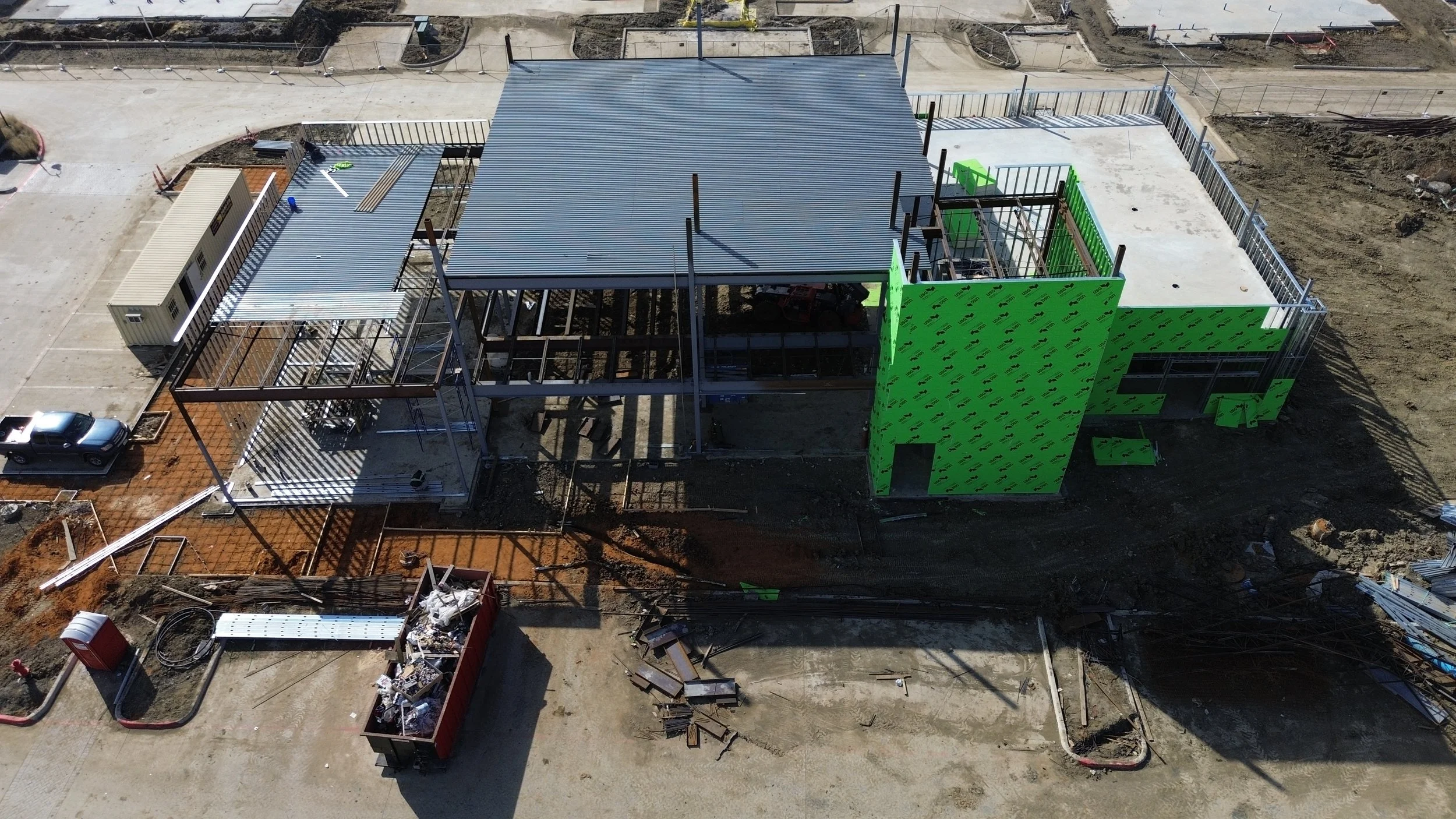 Aerial view of a building under construction with scaffolding, metal framework, and a partially completed roof, surrounded by construction materials and a truck.