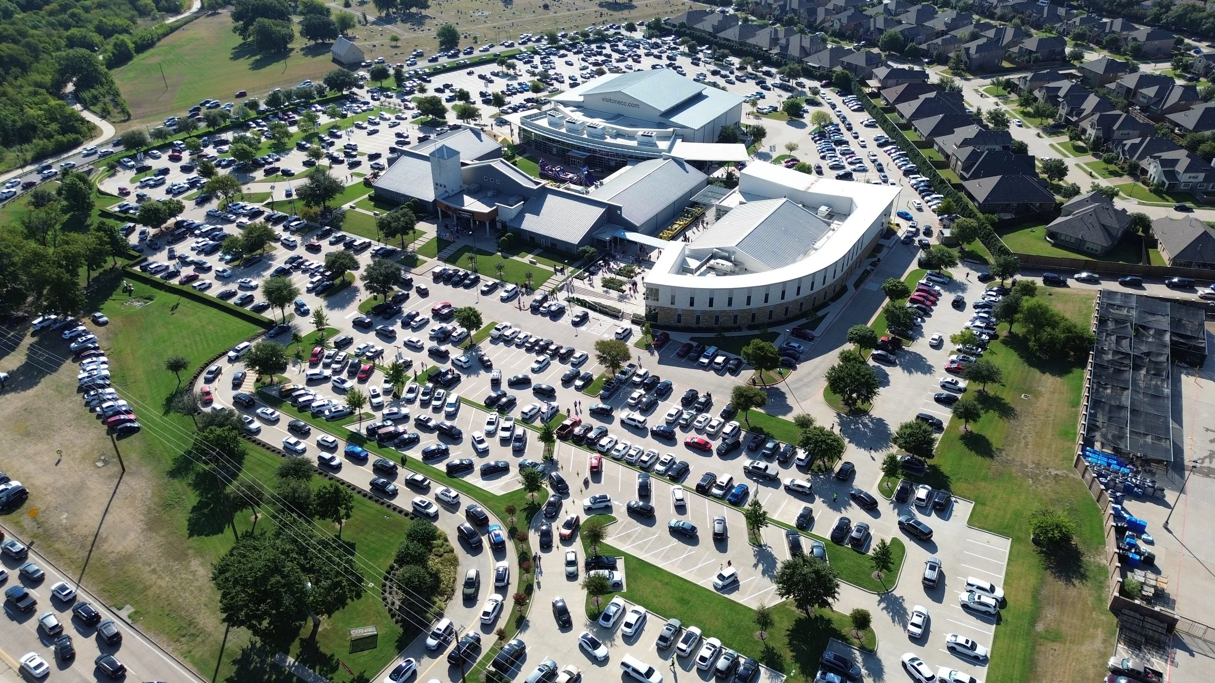 Aerial view of a large parking lot filled with cars, surrounding a modern building complex in a suburban neighborhood. Adjacent roads and houses are visible.