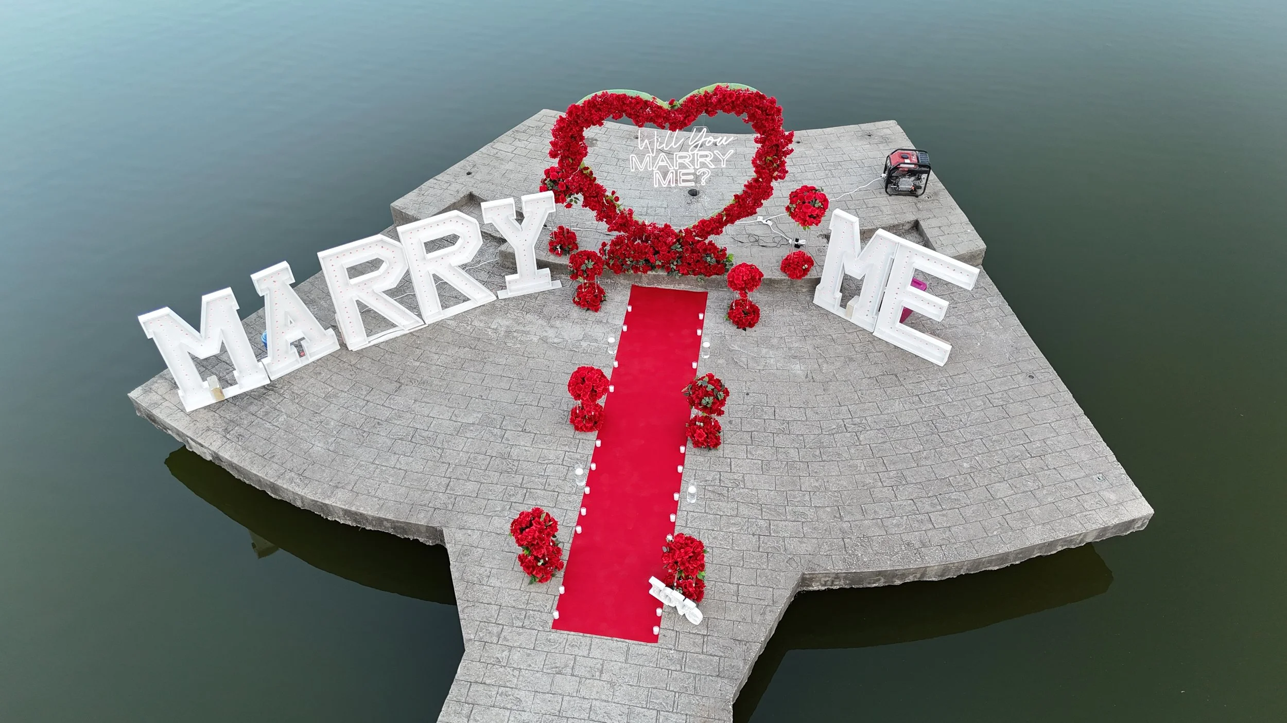 Aerial view of proposal setup with 'Marry Me' sign, red heart, red carpet, flowers, by waterfront.