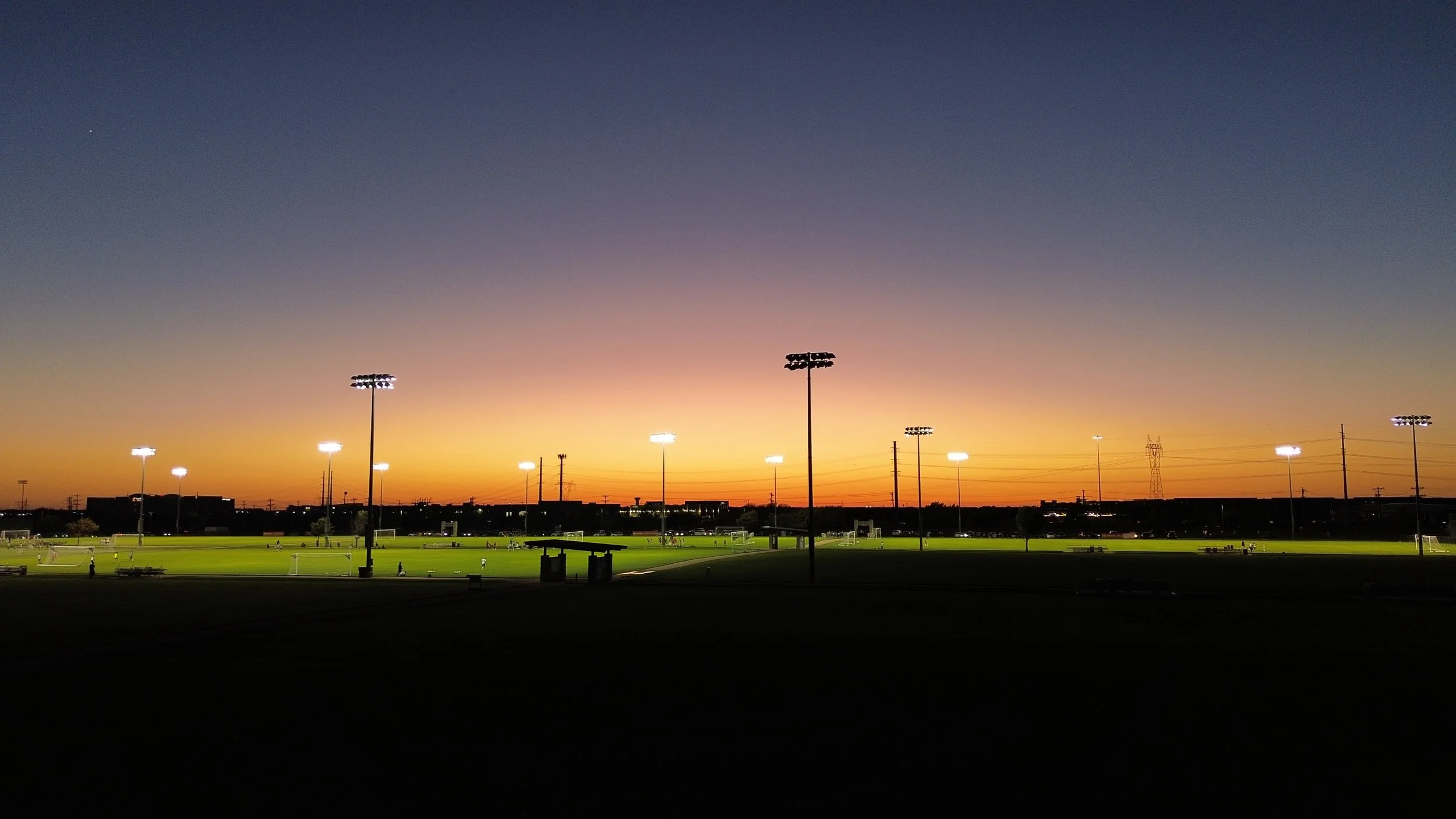 Drone image of sports field at sunset with stadium lights on and orange sky in background.