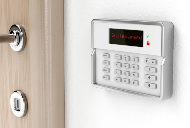 A security alarm keypad mounted on a wall next to a light-colored door, with the screen displaying 'System armed' and icons indicating the system is armed and locked.