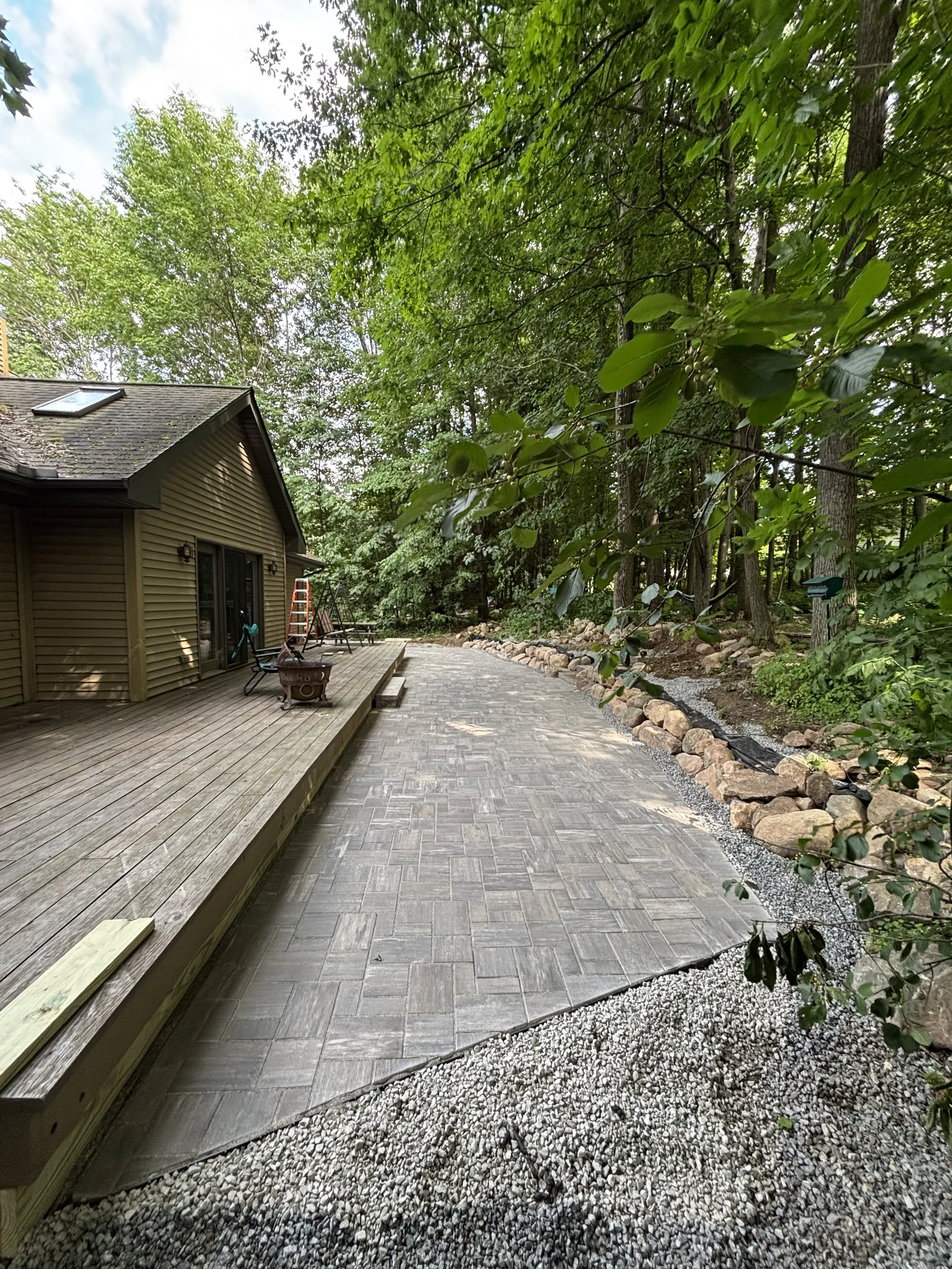 Partially completed backyard patio with a wooden deck, concrete pavers, gravel border, and surrounding trees.