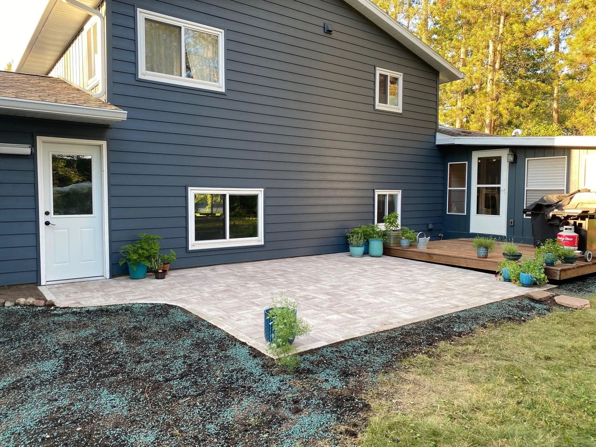 Backyard patio with planted pots, potted plants, and a grill, adjacent to a blue house with multiple windows and a small deck.