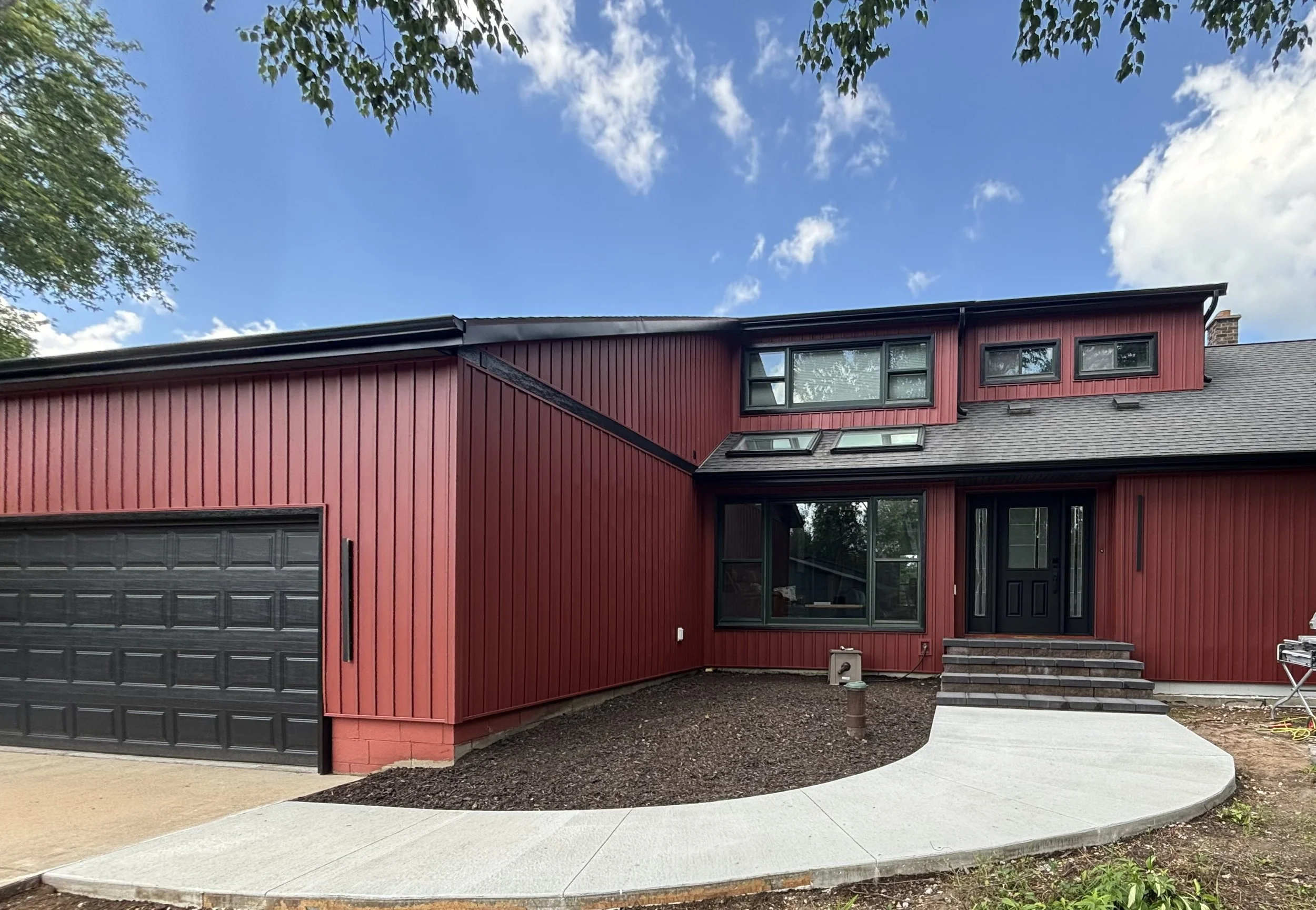 Newly constructed modern two-story house with red vertical siding, black window frames, black front door, concrete walkway, and a gravel landscape area under a partly cloudy sky.