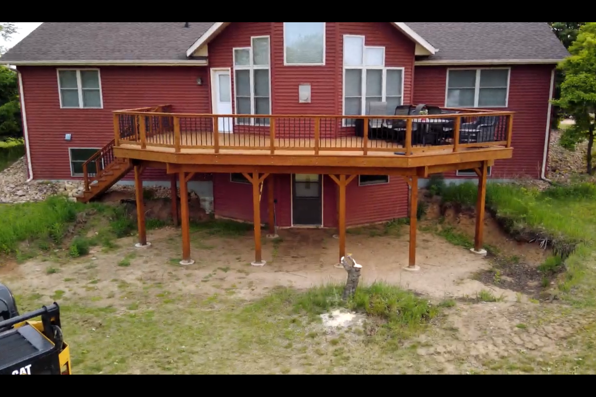 Red two-story house with a wooden deck and stairs, surrounded by green grass and trees.