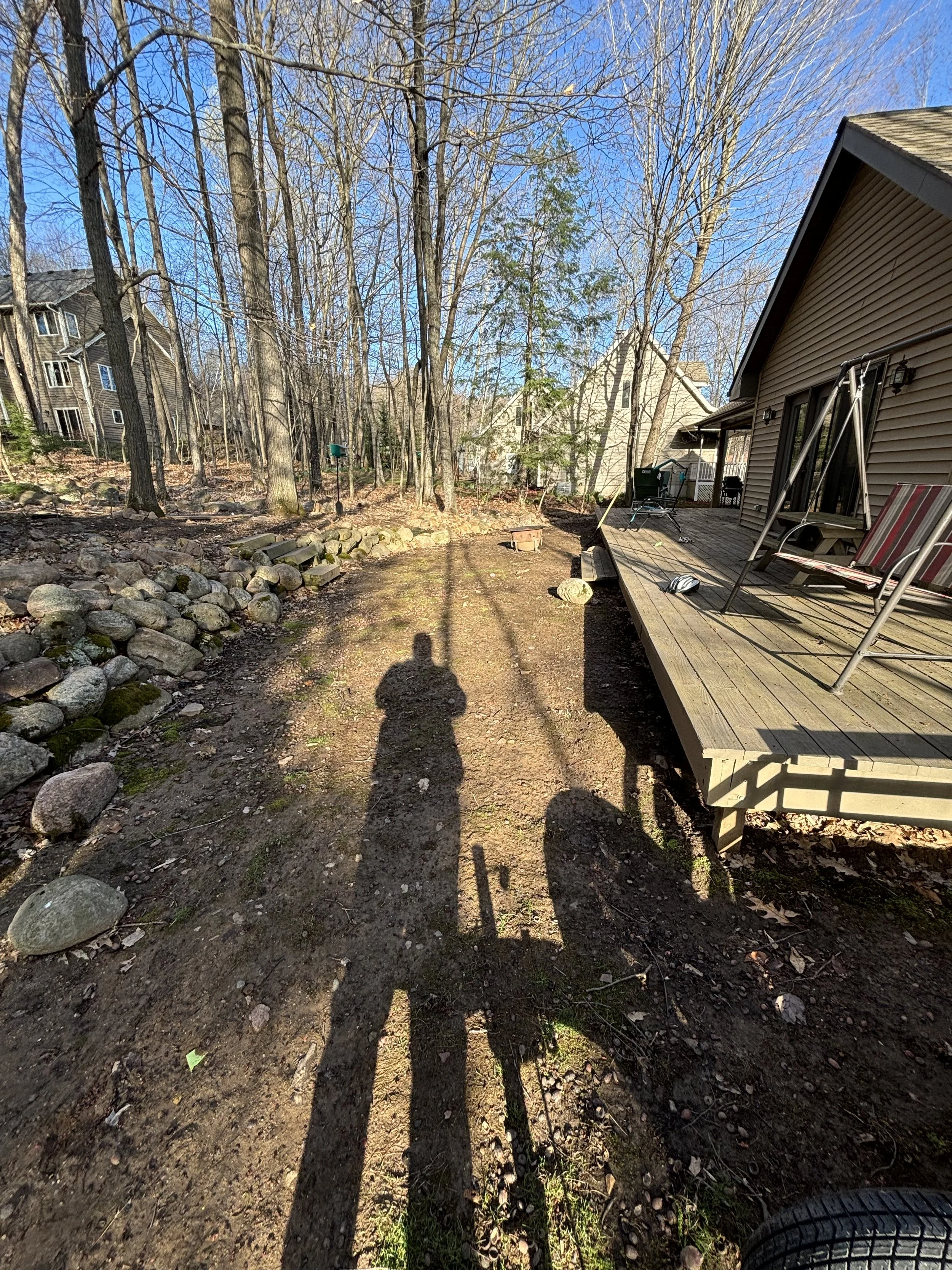 Backyard with trees, a wooden deck with chairs, and a dirt pathway with shadows of a person and bicycle in sunlight.