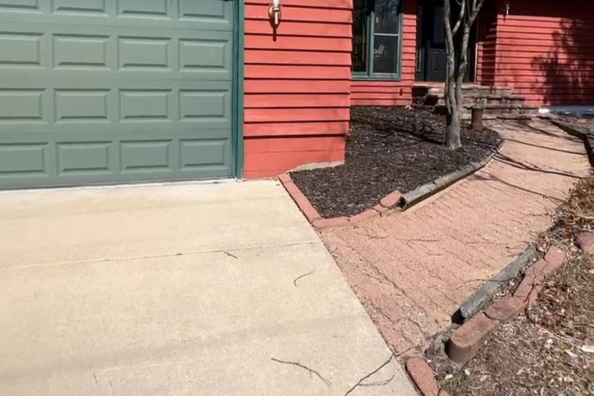A view of a house corner with a green garage door, red siding, a small tree, a light fixture, and a sidewalk made of bricks.