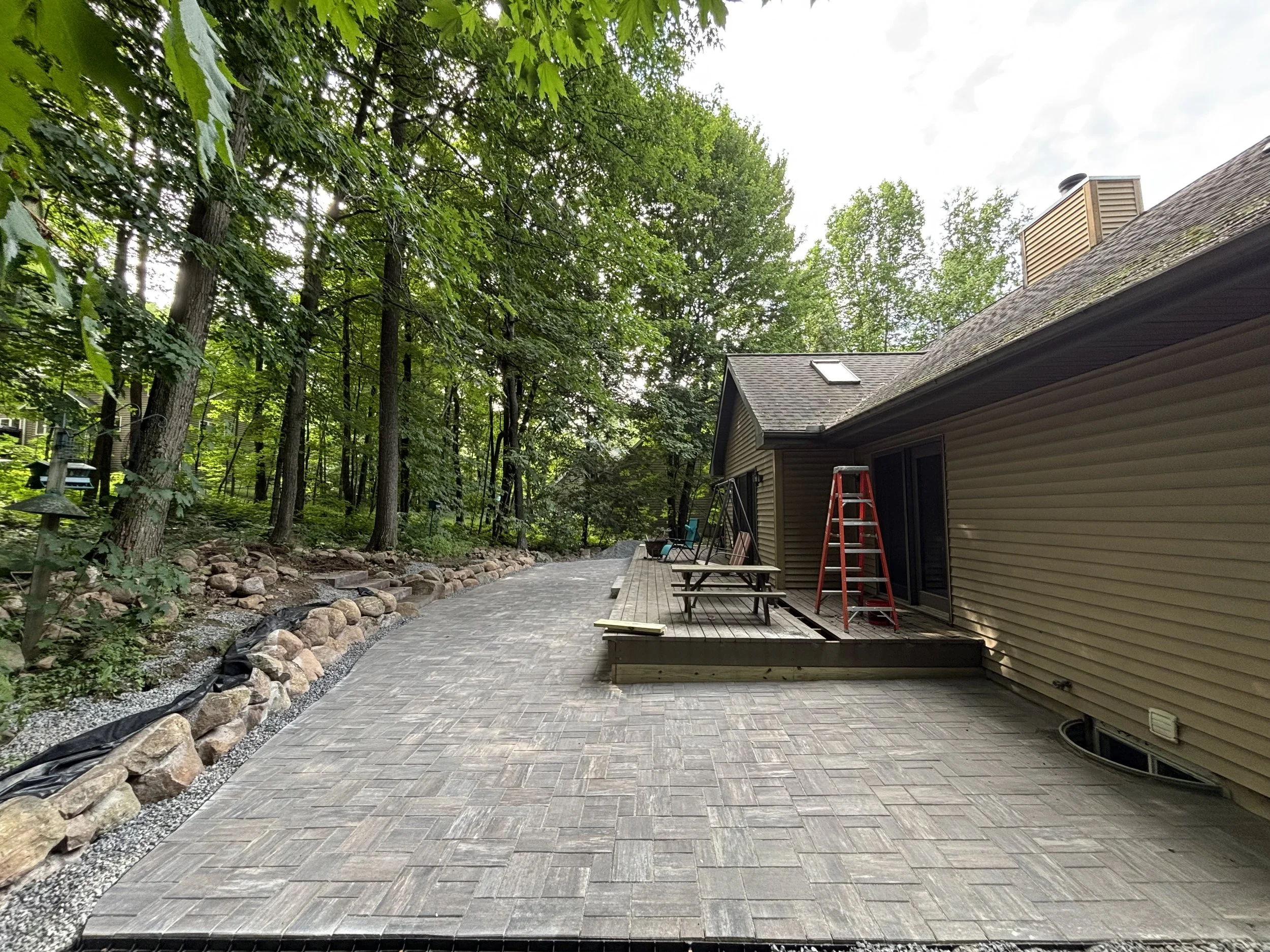 Newly paved patio with a wooden deck, outdoor furniture, and red ladder, next to a house with a sloped roof and surrounded by trees.
