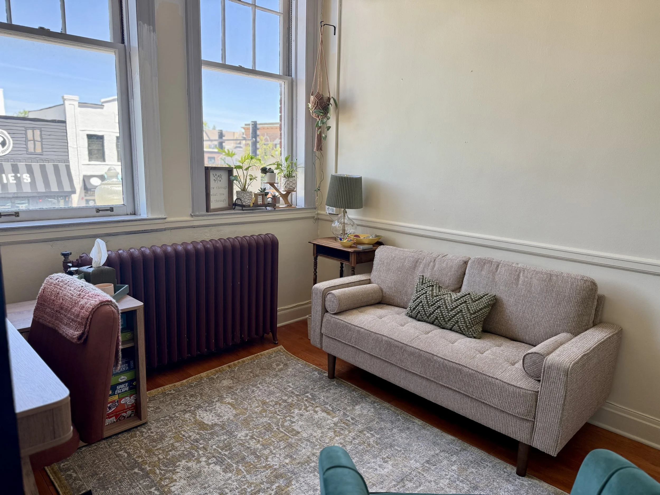 A cozy living room with a beige sofa, a small wooden side table with a lamp, and decorative pillows. There are tall windows with gardens outside, a small framed quote, and plants on the windowsill. A patterned rug covers the wooden floor.