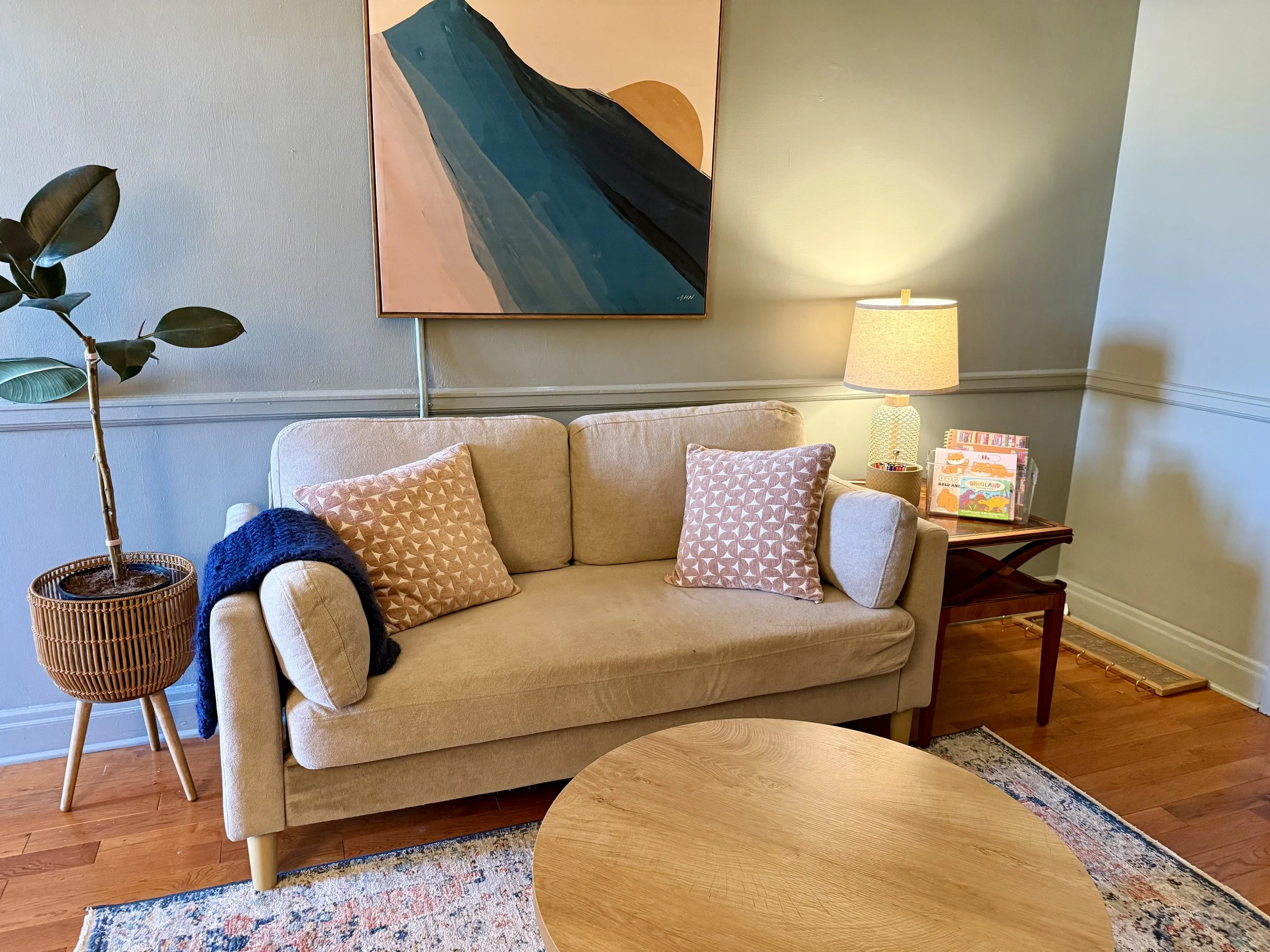 A living room corner features a beige sofa with two pink patterned pillows, a black blanket, a wooden coffee table, a tall potted plant, a turquoise abstract landscape painting, and a table lamp with a textured beige shade. There are children's books on a side table, and hardwood flooring with a patterned rug.