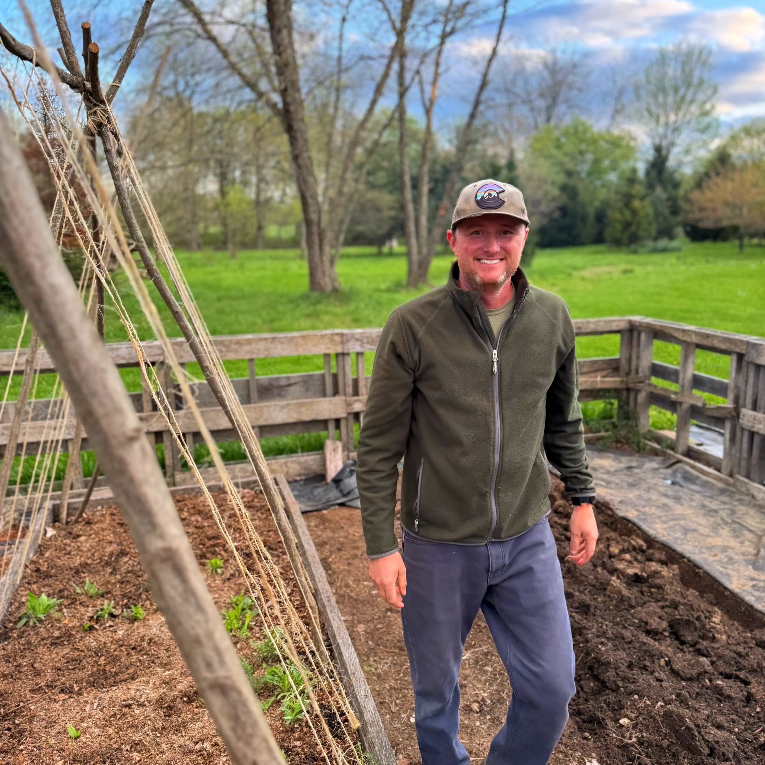 A man in a green jacket and baseball cap smiling outdoors in a garden with freshly tilled soil and a wooden fence, trees, and green grass in the background.