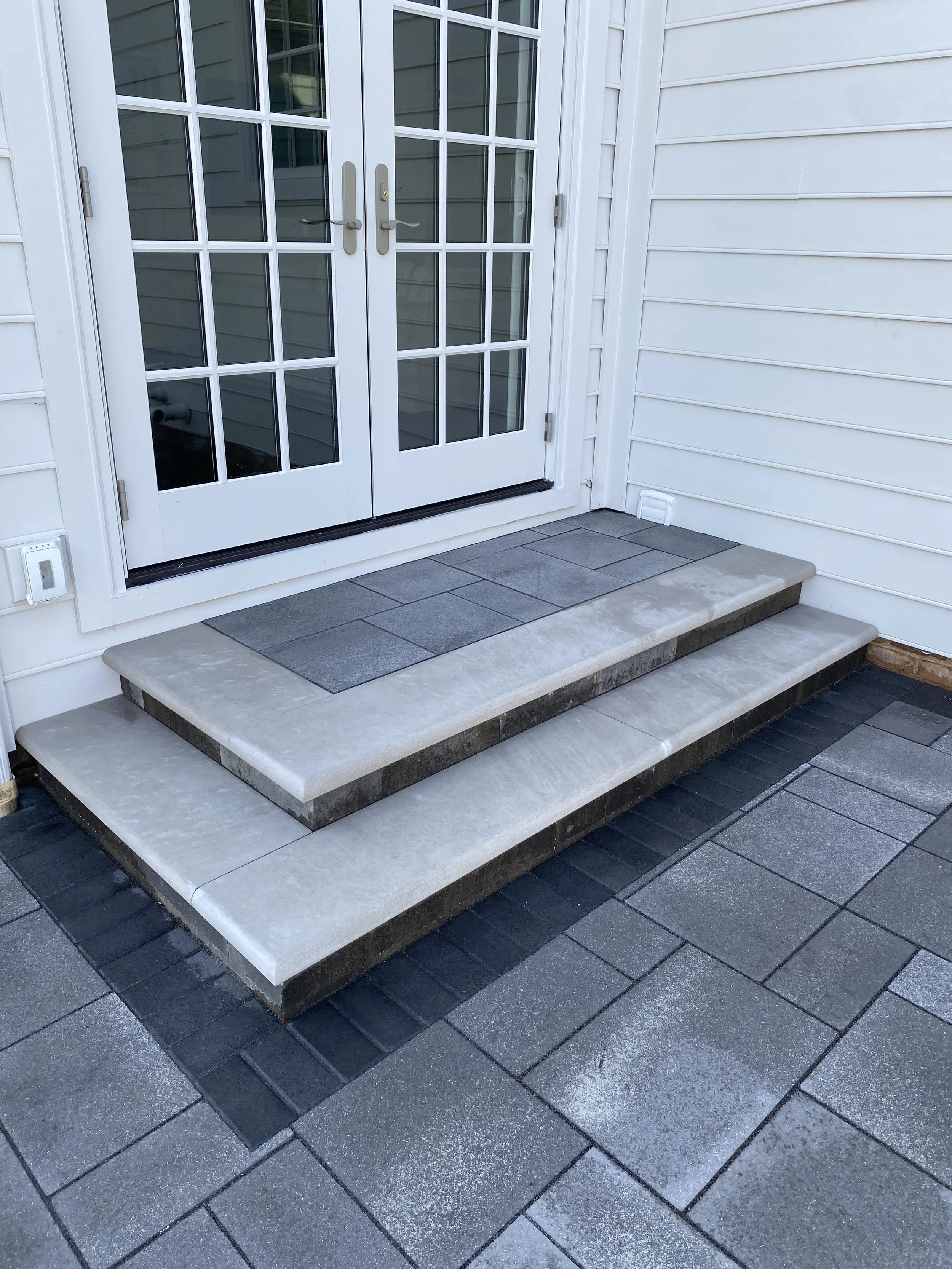 Indiana limestone steps and Unilock Umbriano paver patio leading to a set of white double glass doors with a metal handle, attached to a house with white siding.