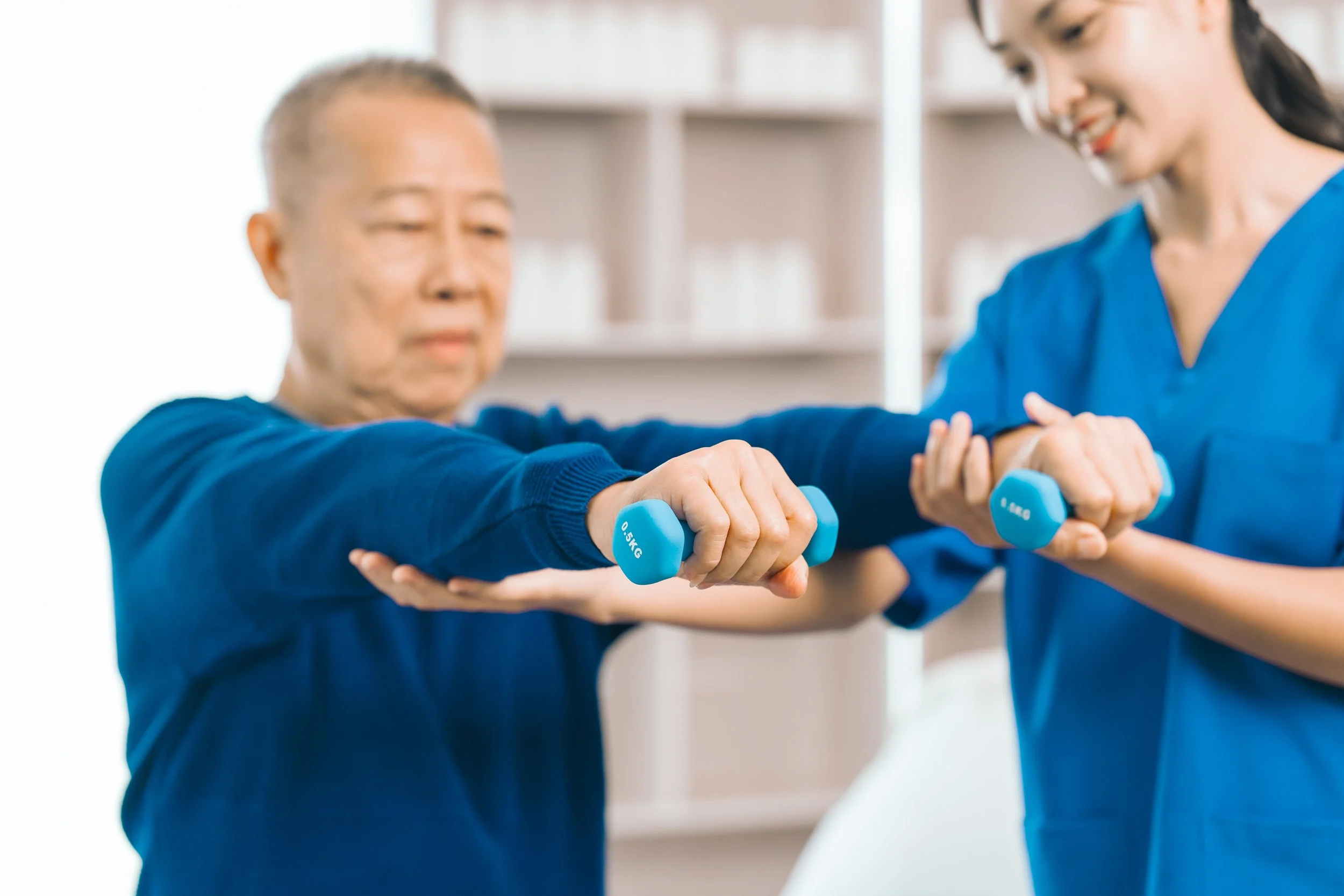 A healthcare worker assists an elderly woman in a physical therapy exercise using light blue dumbbells in a clinical setting.