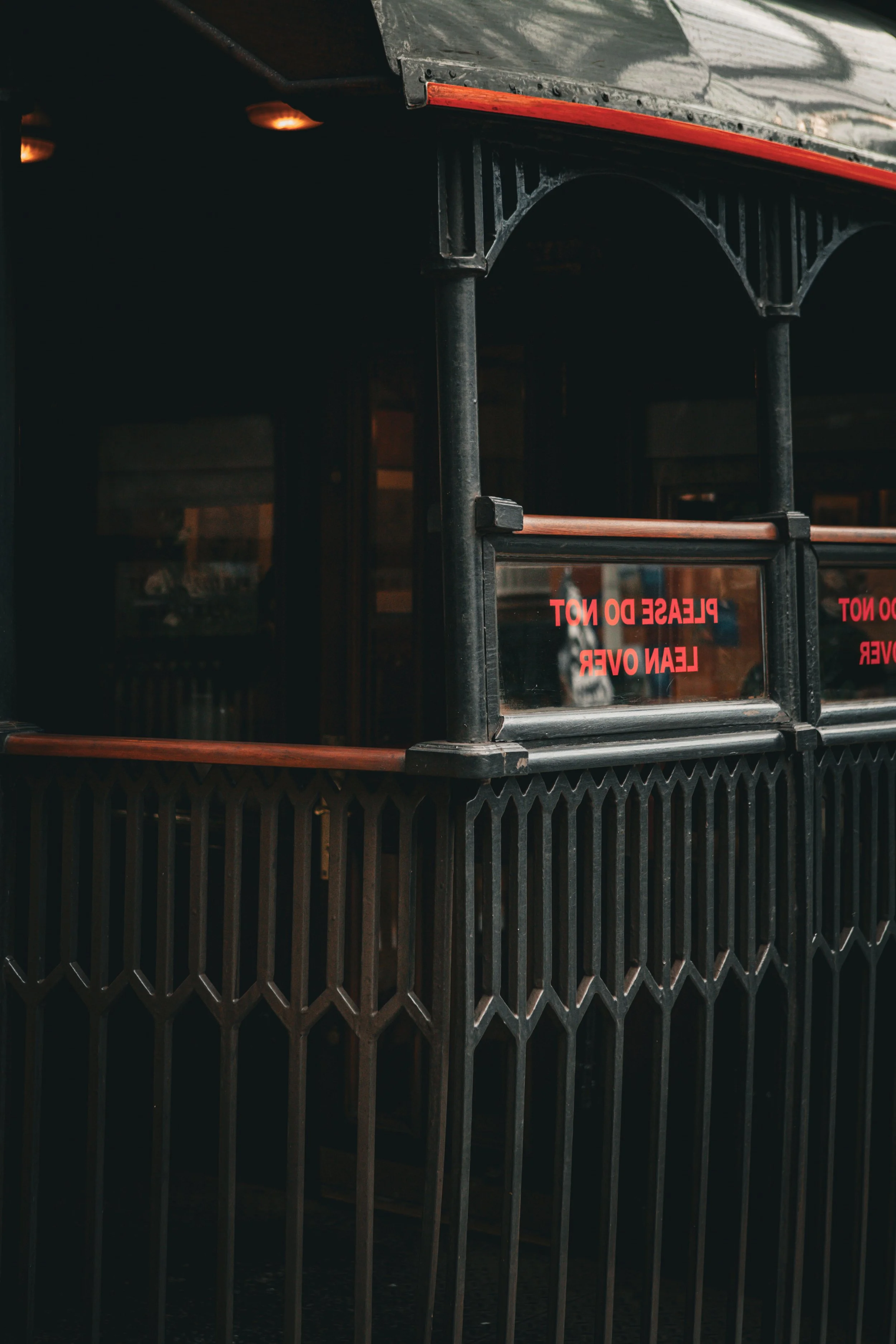 Close-up of a dark protective barrier with some red-orange accents and a glass window with a red sign reading 'PLEASE DO NOT LEAN OVER' reflected in it.