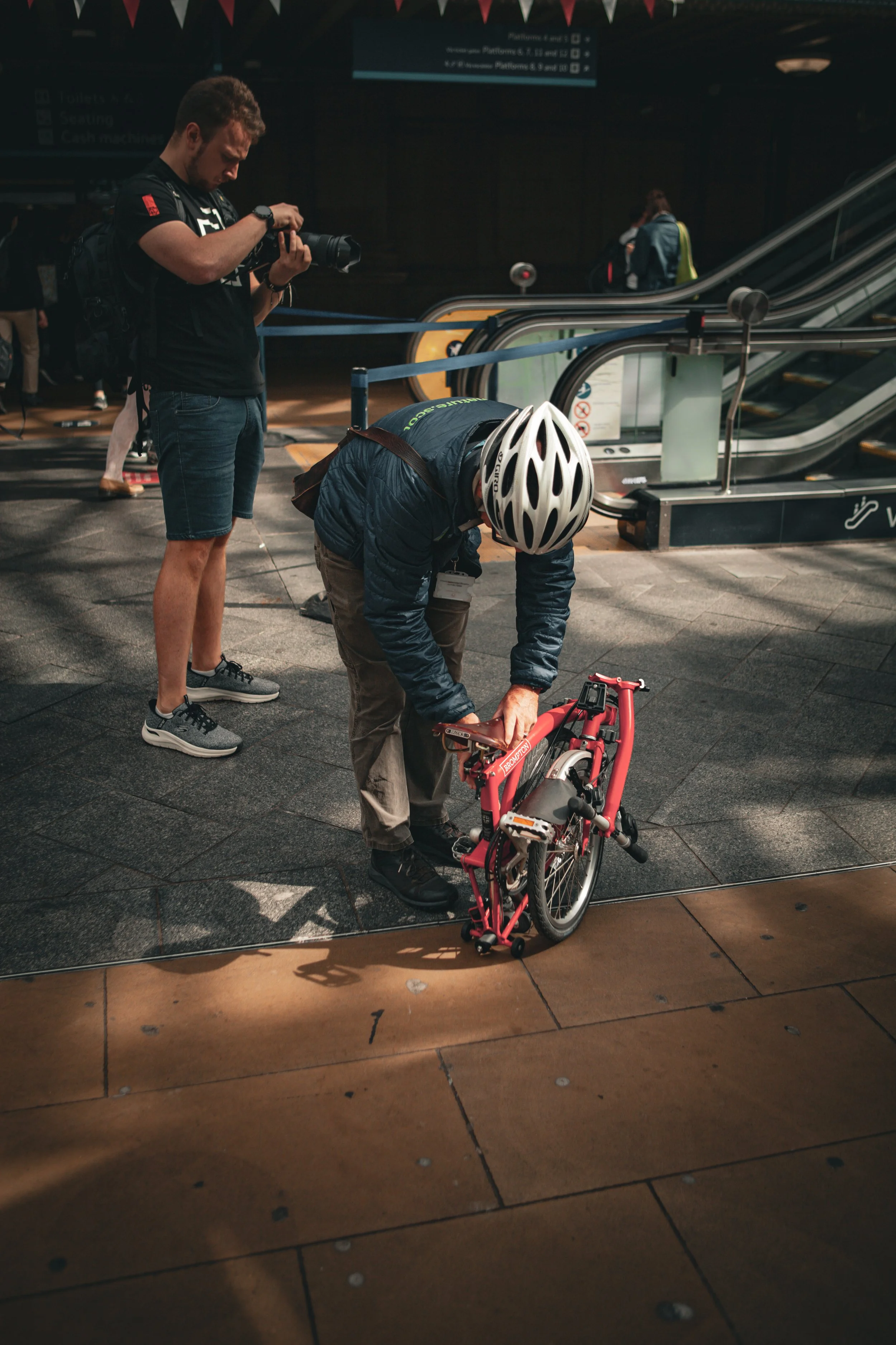 A man with a bicycle and a helmet, bending down to close or open the bike's folding mechanism, standing on a tiled floor in an indoor transit station. A photographer is taking a picture of him. In the background, there are escalators and a few people