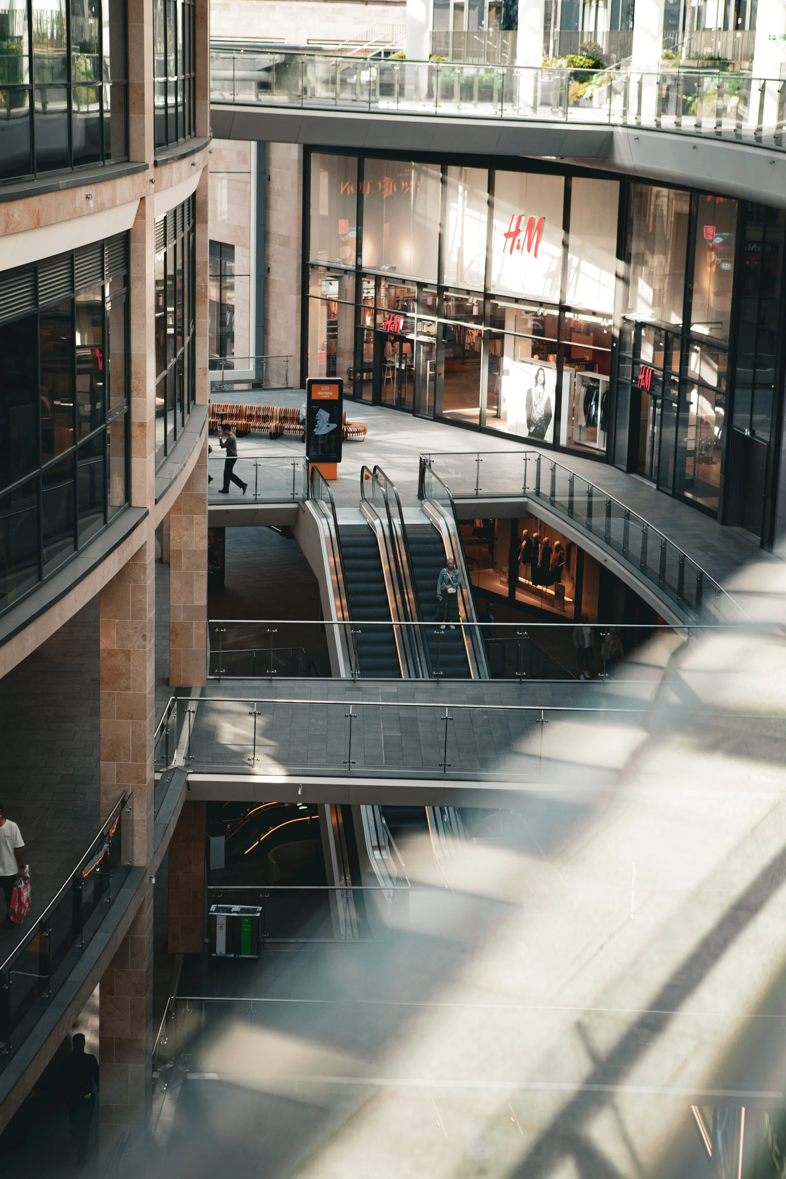 Interior of a multi-level shopping mall with escalators, glass railings, and a visible H&M store entrance.