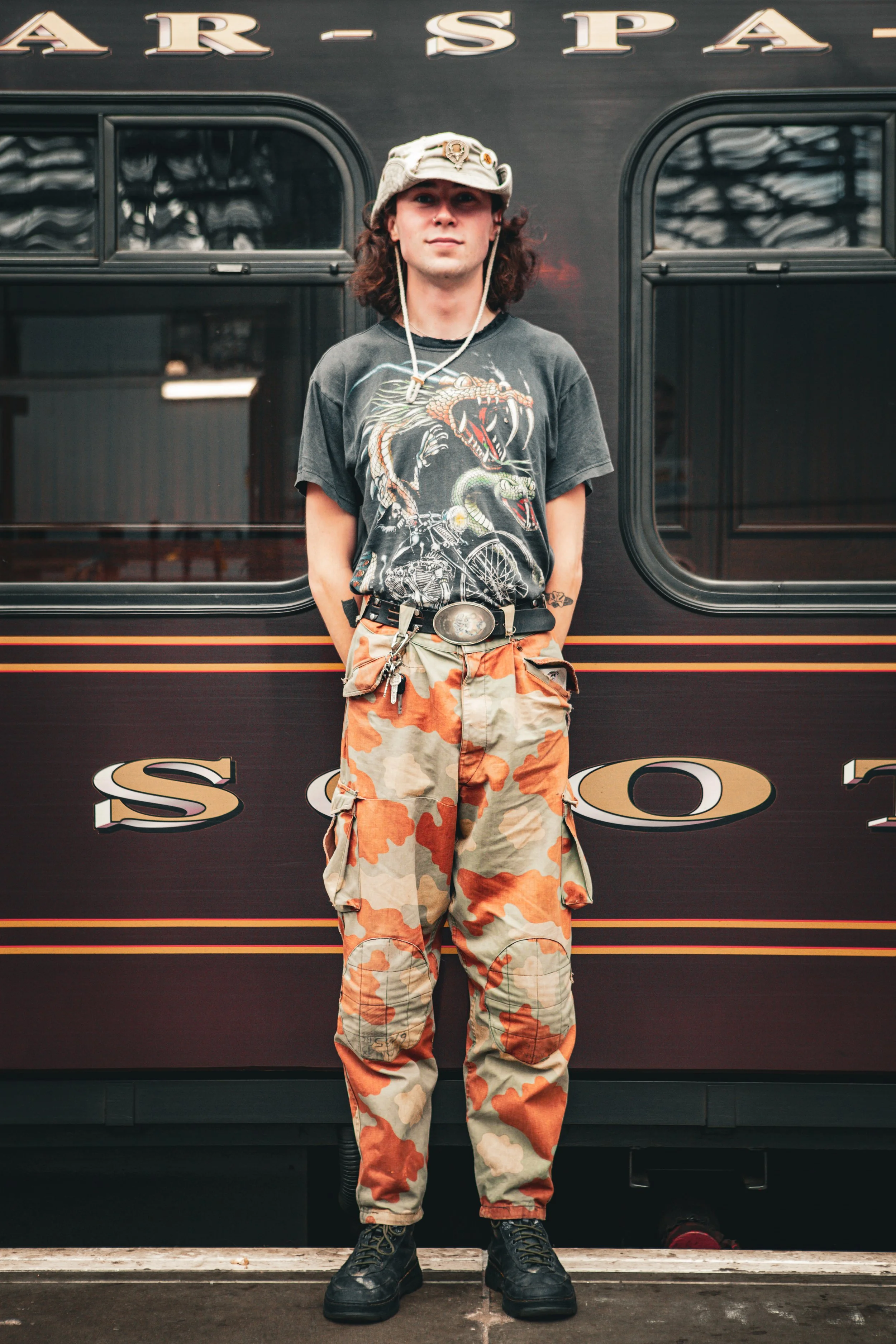 Young man standing in front of a train car, wearing a hat, a graphic t-shirt, camouflage pants, and black boots.