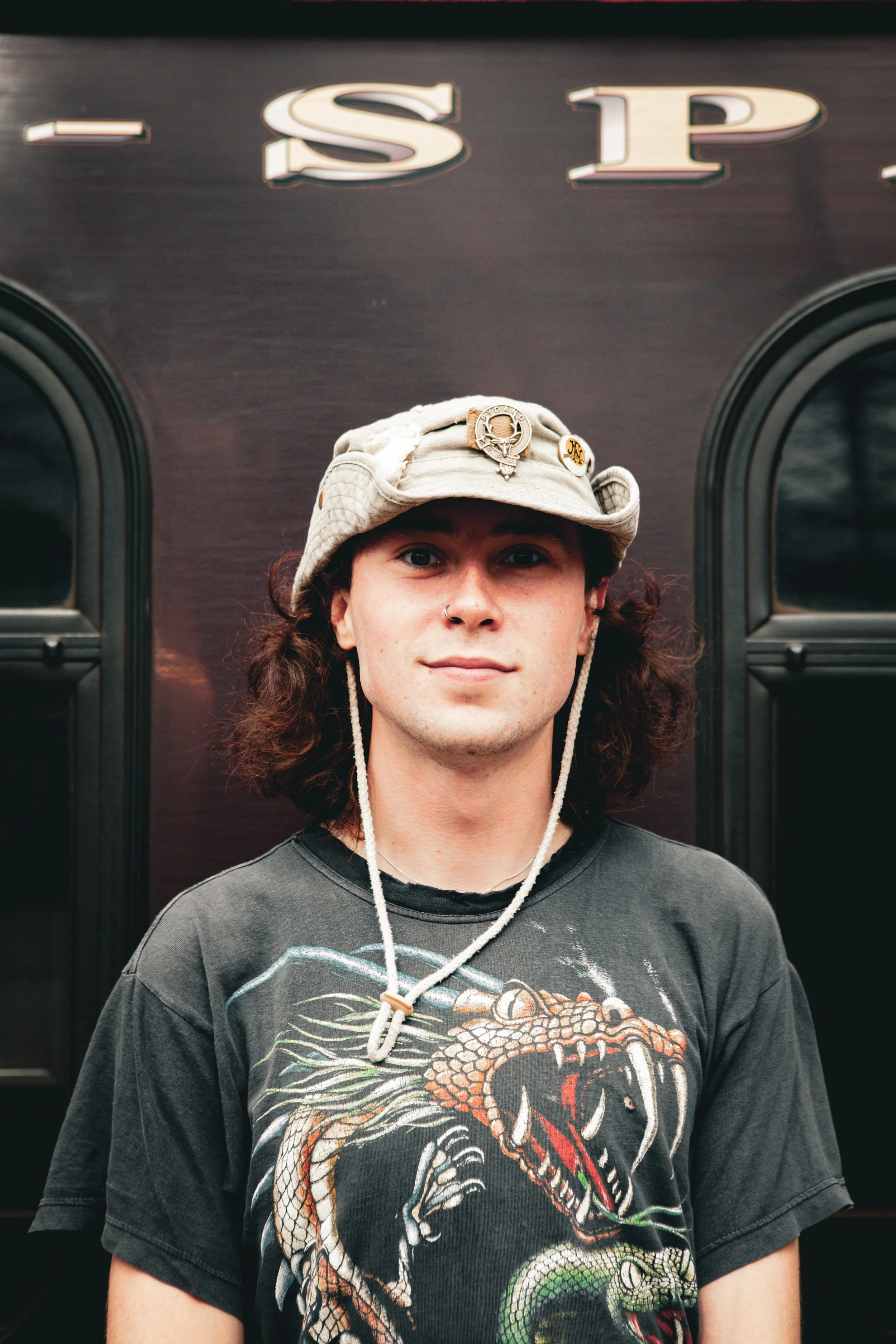 Young man with curly hair wearing a beige hat with pins, a nose ring, and a black T-shirt featuring a dragon illustration, standing in front of a dark vehicle with the letters 'LSP' on it.