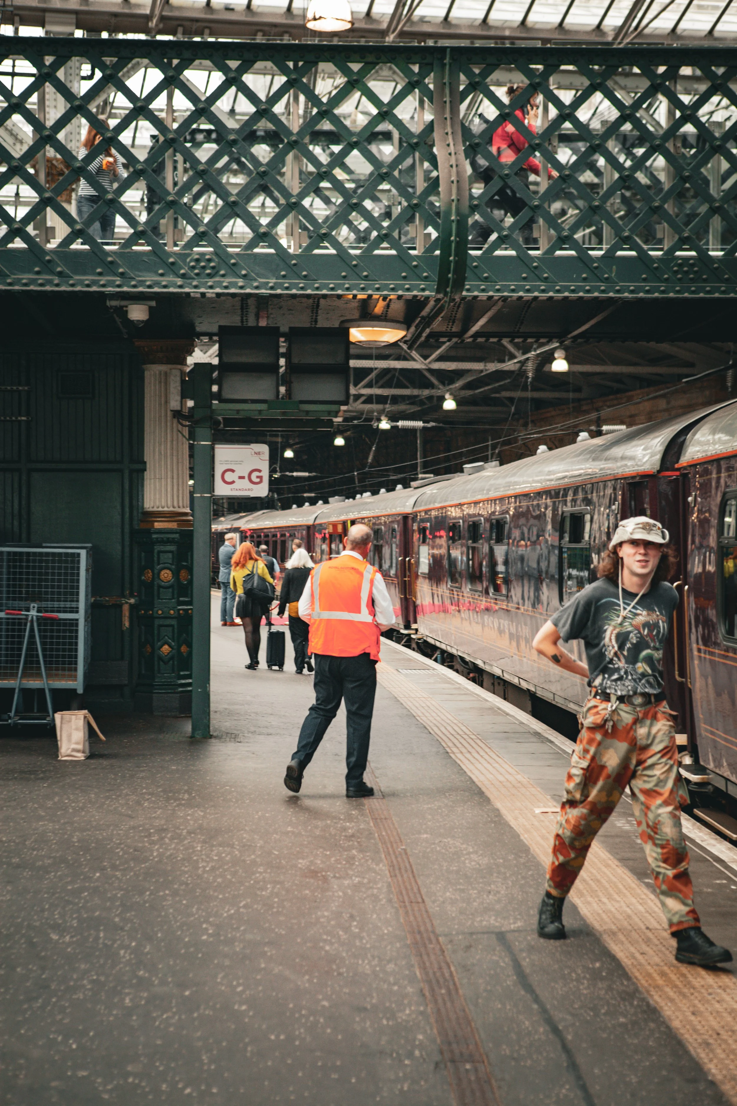 People boarding and disembarking a vintage train at a train station. The station has a platform with a bridge overhead, with some individuals carrying luggage.
