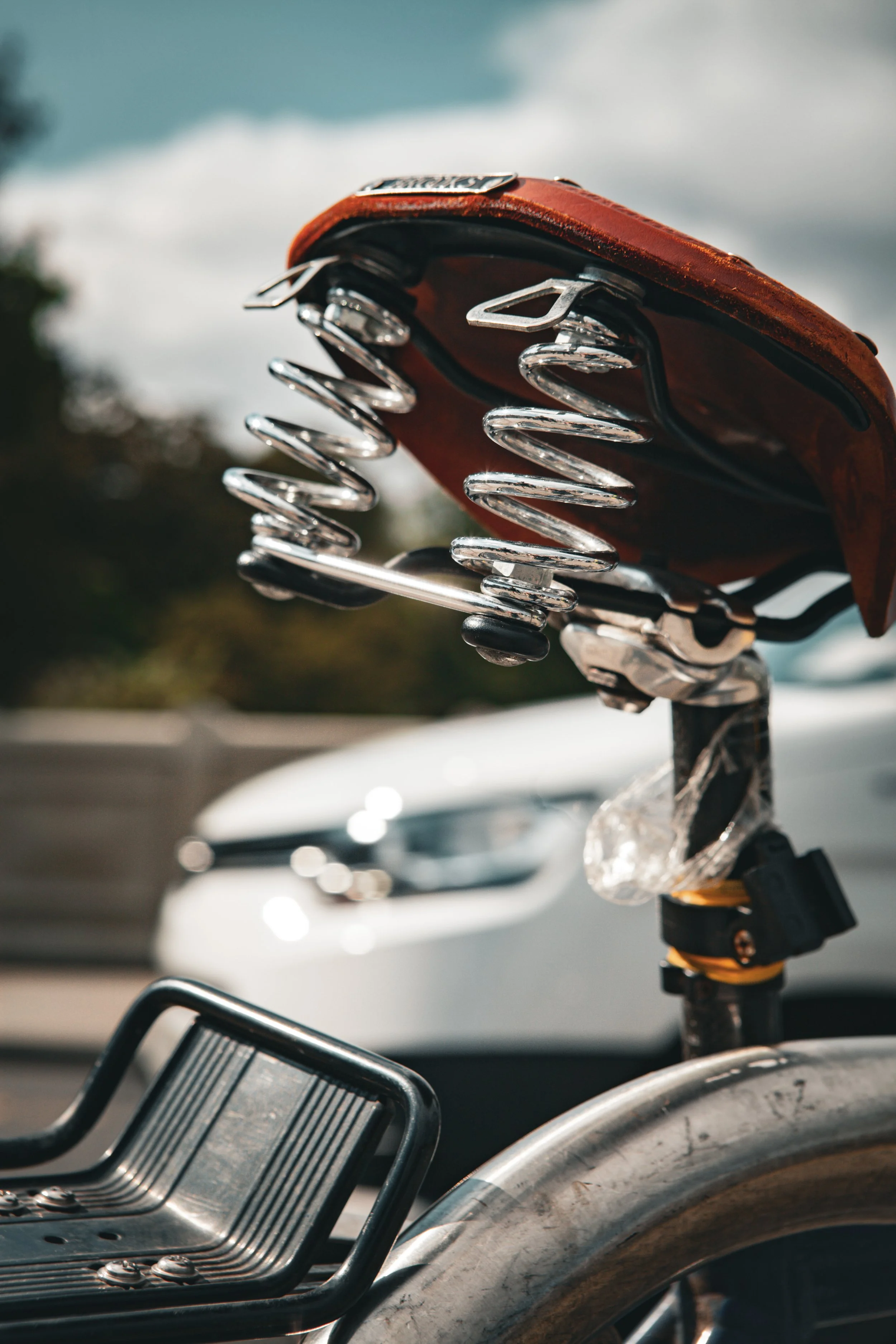 Close-up of a bicycle seat with a leather covering, metal springs, and a plastic reflector, with an out-of-focus white car and a cloudy sky in the background.