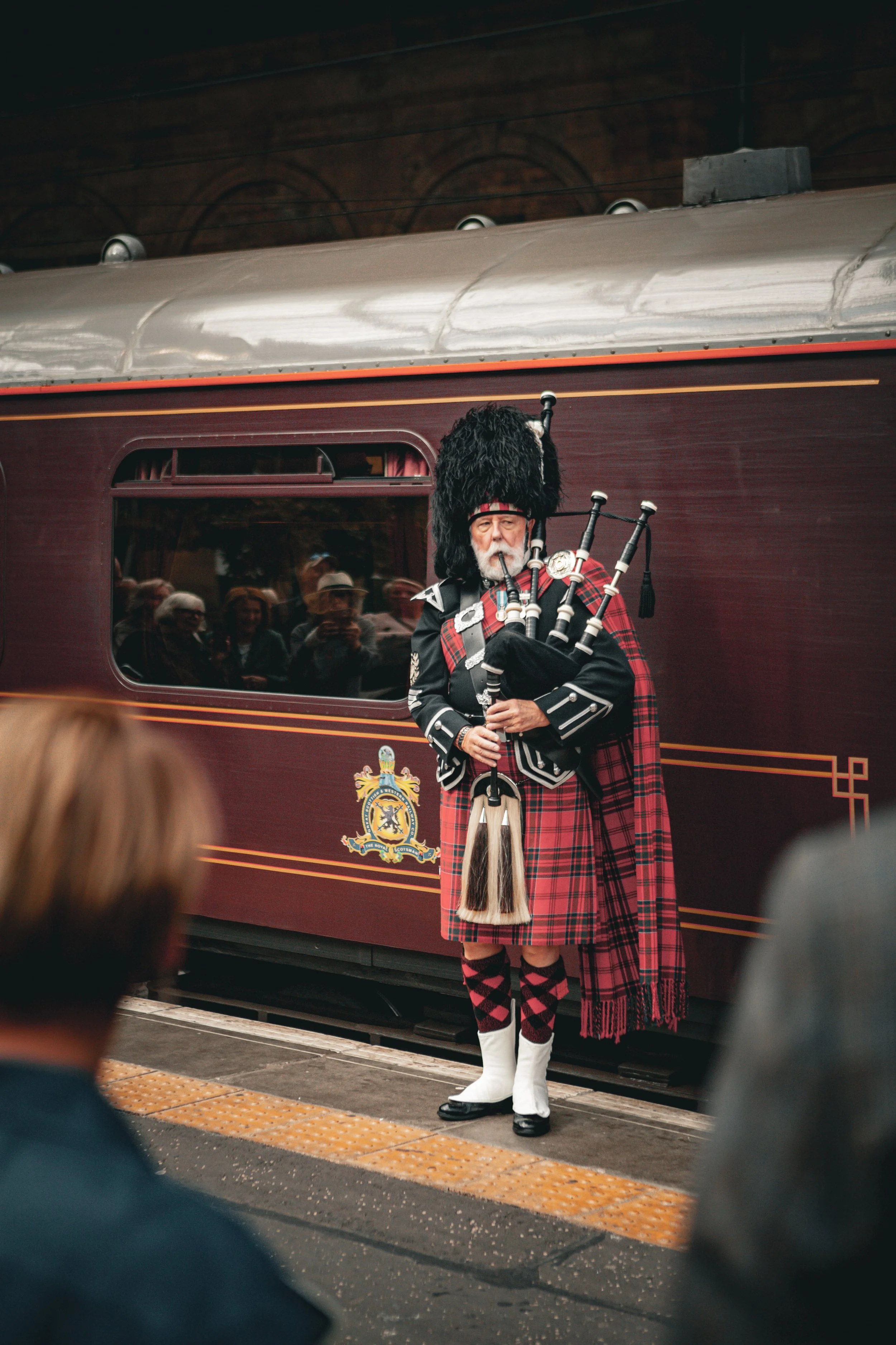 A man dressed in traditional Scottish attire playing bagpipes in front of a vintage train carriage.
