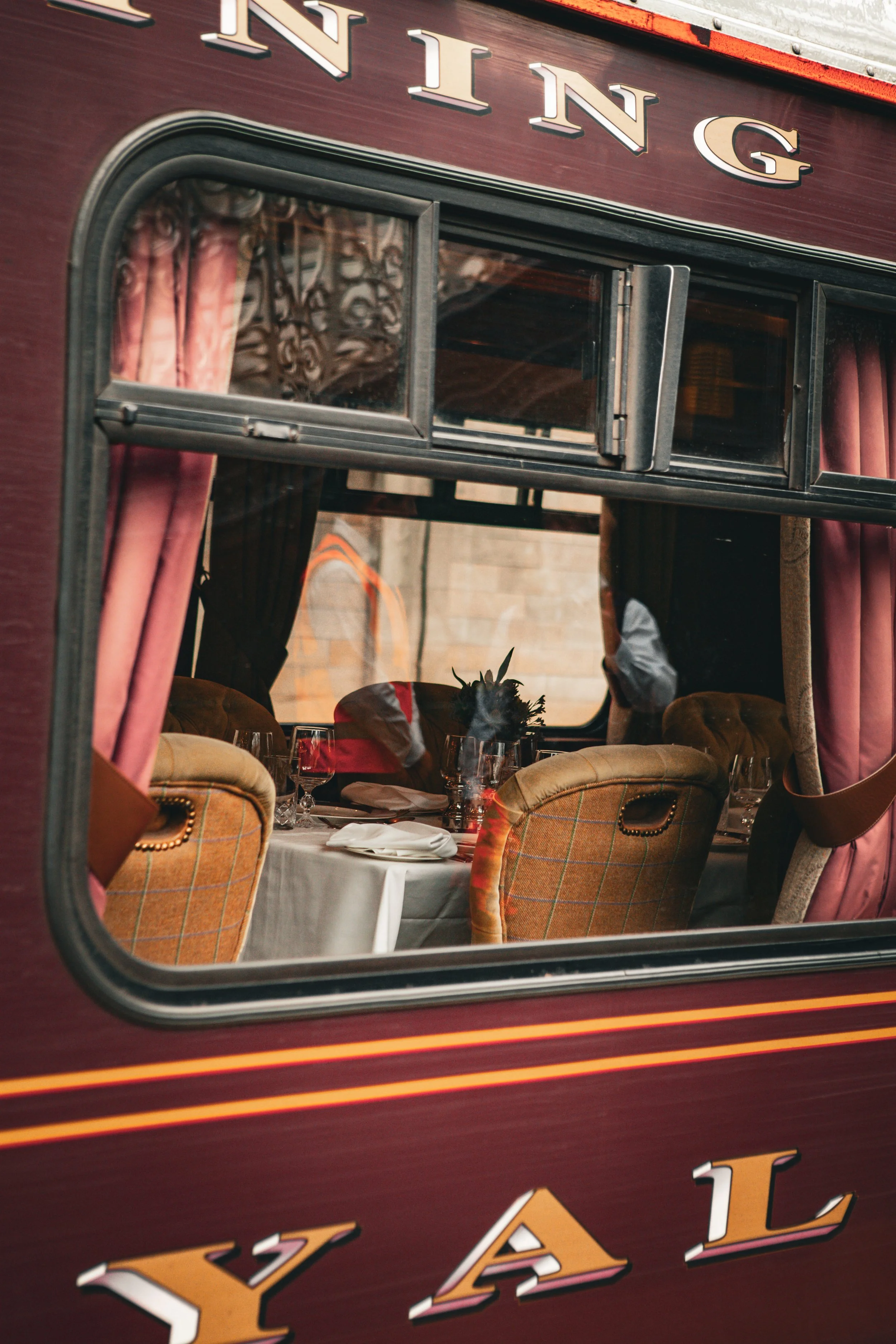 View through a train window showing a dining table set with glasses, plates, and napkins, with velvet curtains and vintage-style chairs inside the train.
