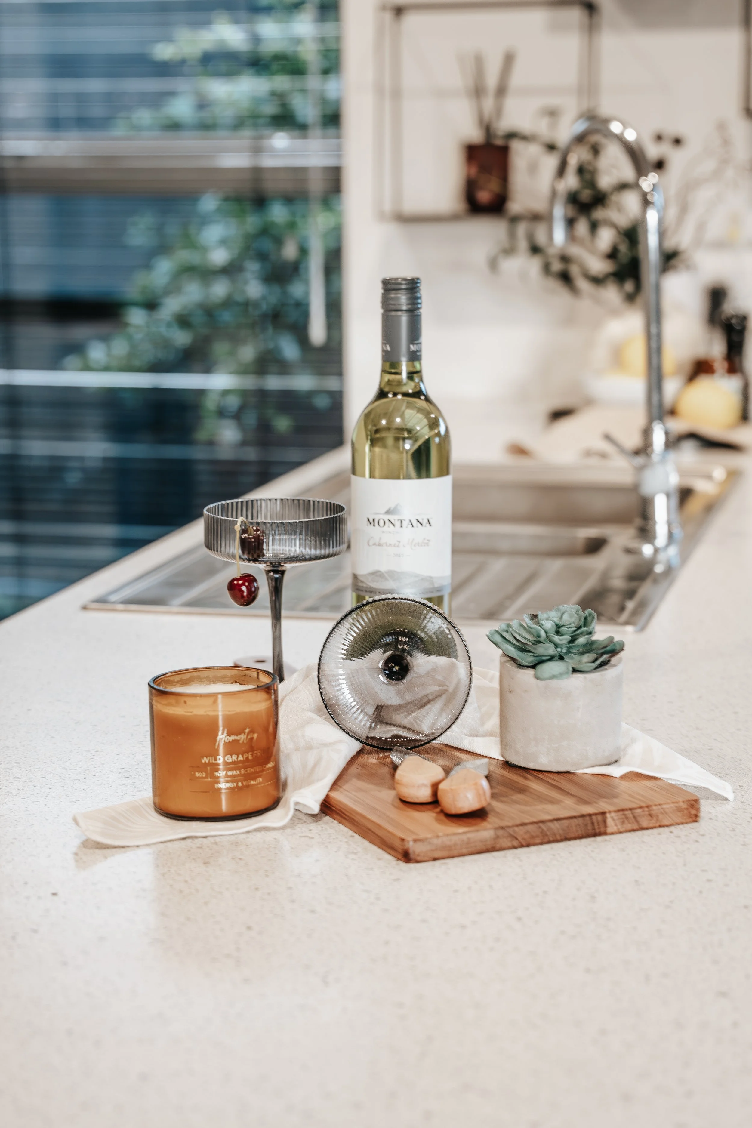 Kitchen countertop with a bottle of white wine, a green succulent plant, a wooden cutting board with pink stones, a candle, and glassware, near a sink and window.
