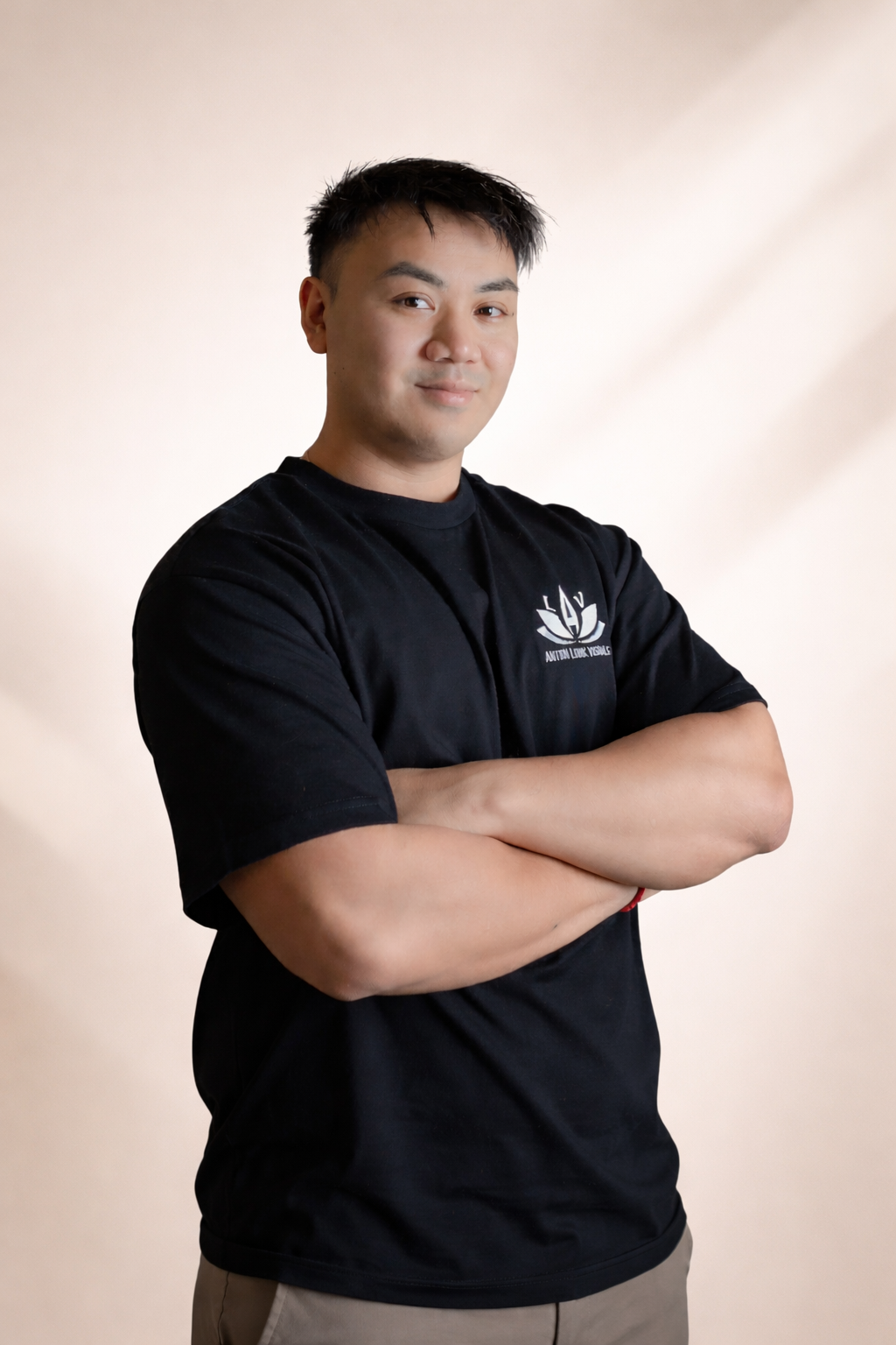 A young man with short black hair standing with arms crossed, wearing a black t-shirt with a logo on the chest, in front of a beige background.