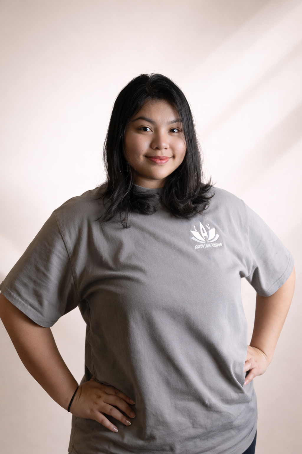 A young woman with dark hair smiling, wearing a gray T-shirt with a logo that says 'Nation Link Visuals', standing with one hand on her hip in front of a neutral background.