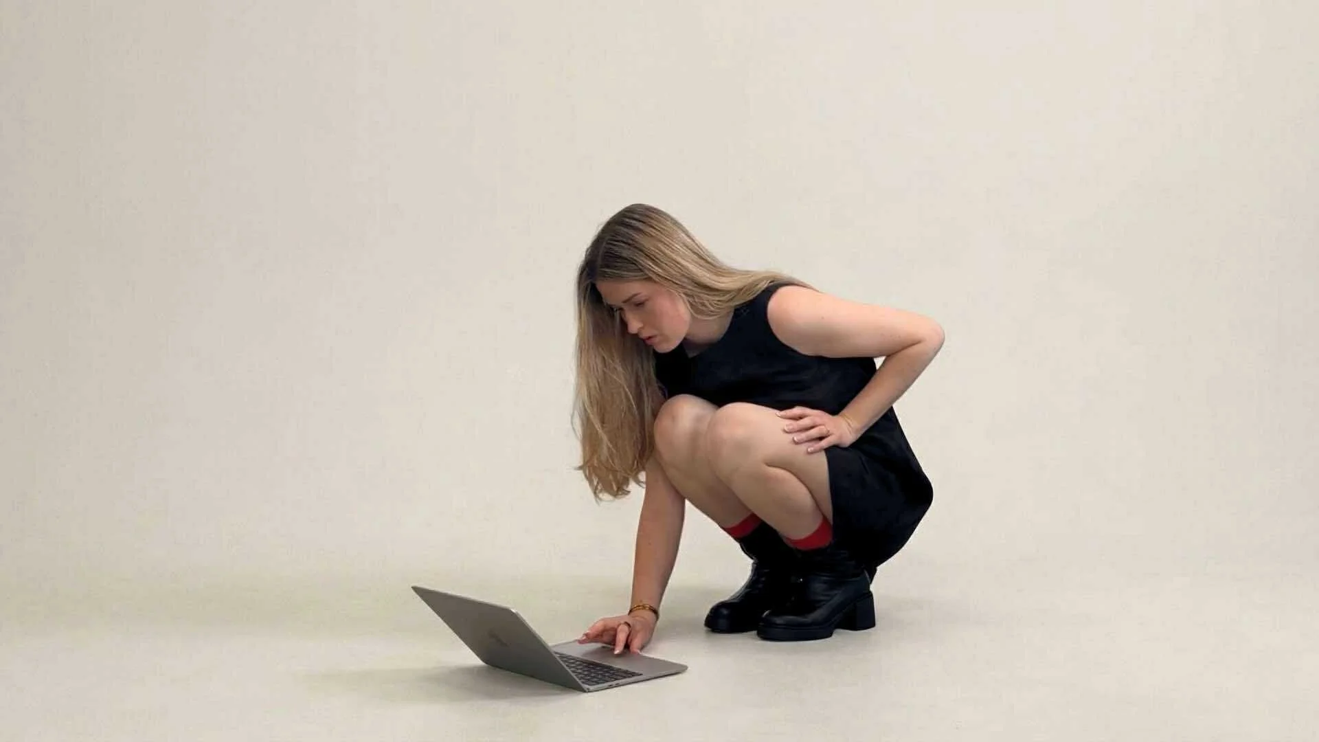 Young woman crouching in black outfit looking at a laptop on the floor against a plain light-colored background.