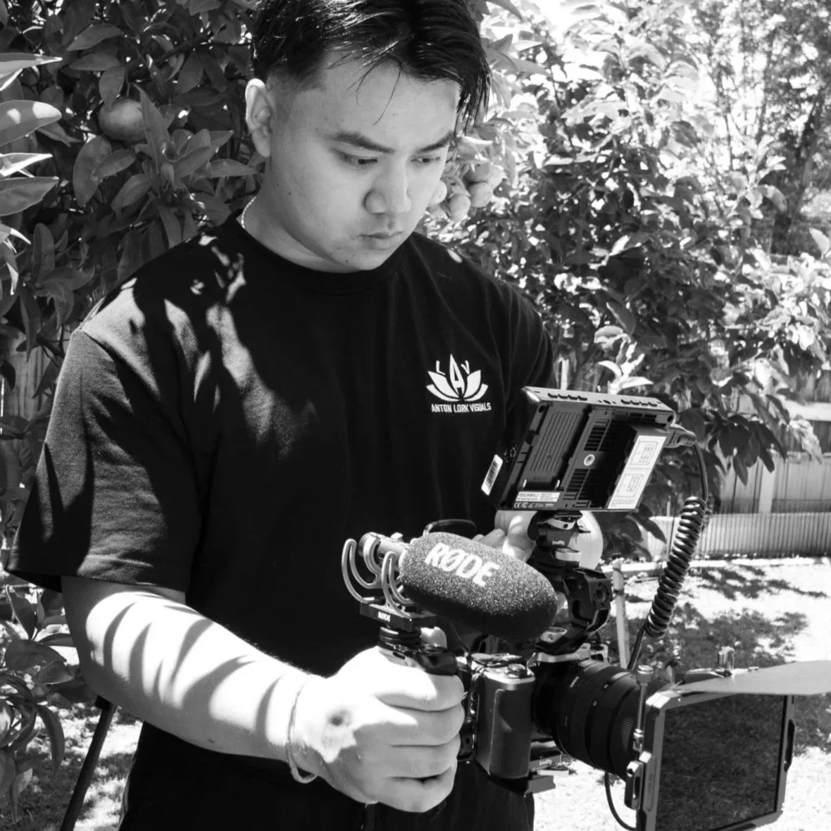 A man in a black t-shirt operating a professional camera with a Rode microphone attached, outdoors among trees.