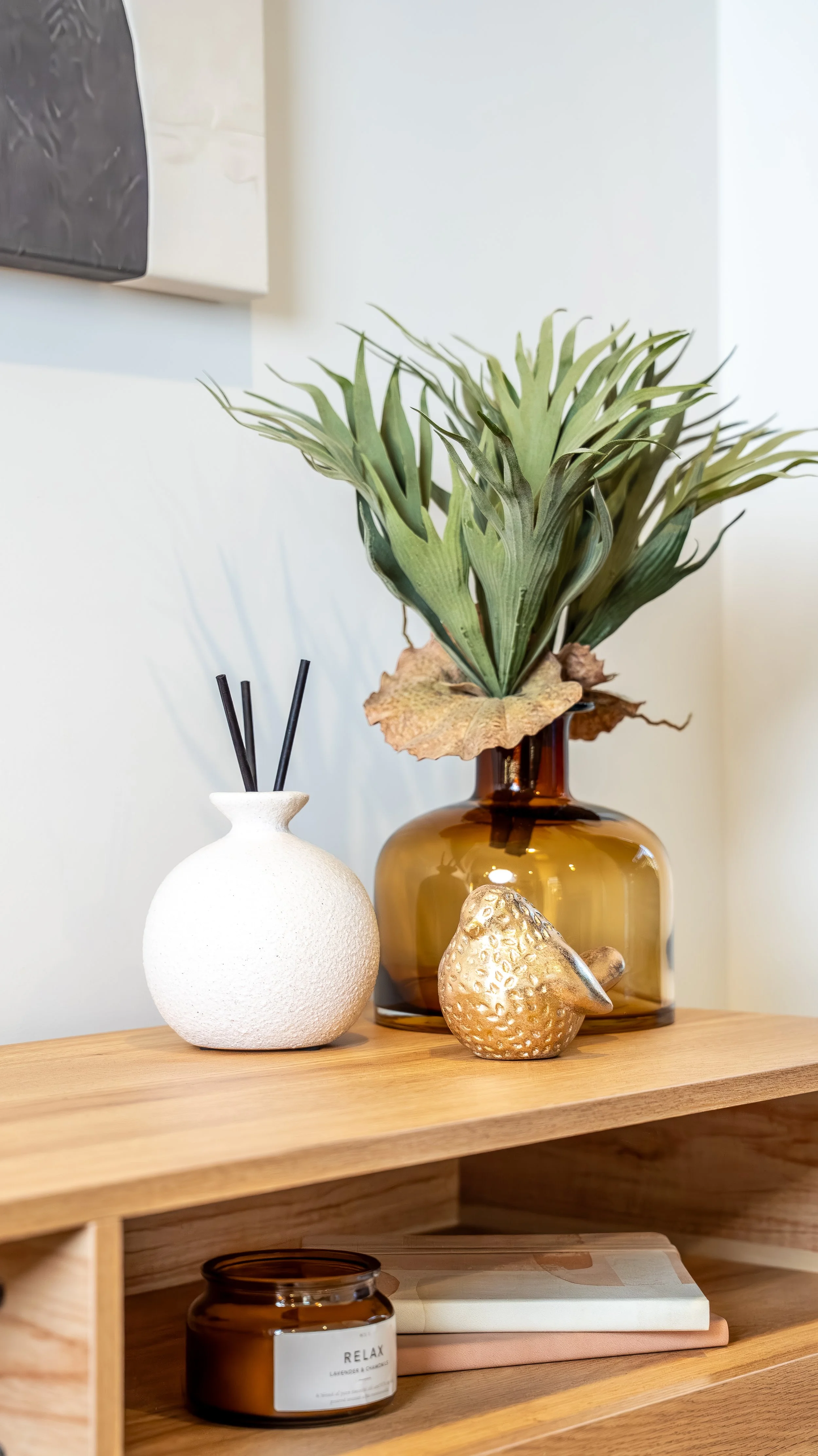 Decorative arrangement on a wooden shelf includes a brown glass vase with green leaves, a white textured sphere-shaped diffuser with black sticks, a gold textured pear ornament, and a small brown candle jar labeled 'RELAX' with a candle inside.