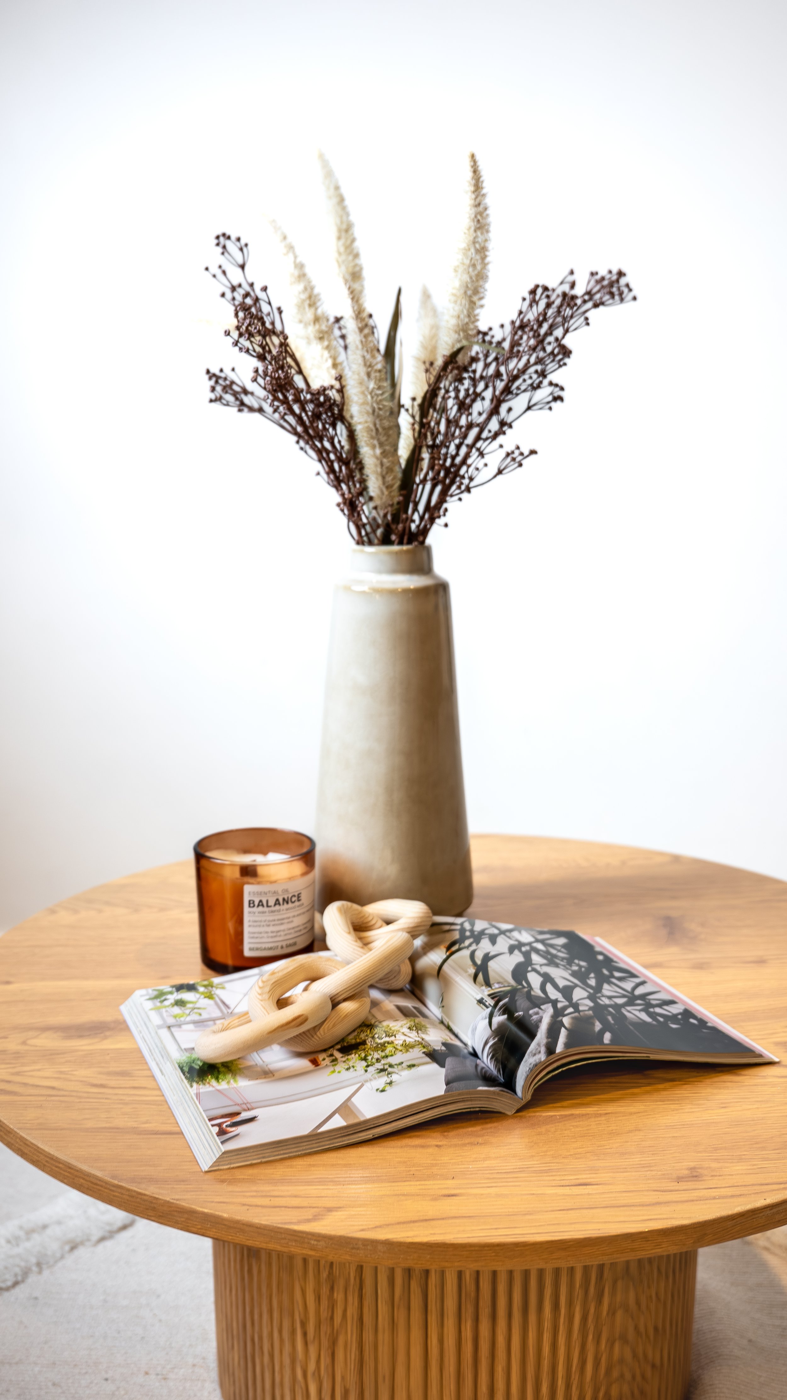 A round wooden table holds an open magazine, a beige chain-link sculpture, a brown candle labeled 'Balance,' and a tall ceramic vase with dried brown and cream flowers.