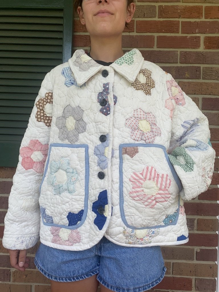 Person wearing a white quilted jacket with colorful patchwork flower patterns and dark blue buttons, standing in front of a brick wall and a green window shutter.
