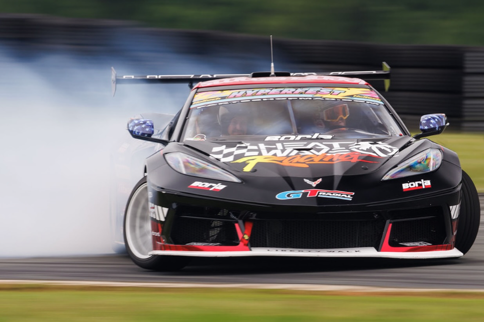 A black race car with a large rear wing, drifting on a racetrack, generating tire smoke. The car has racing decals and sponsorships.