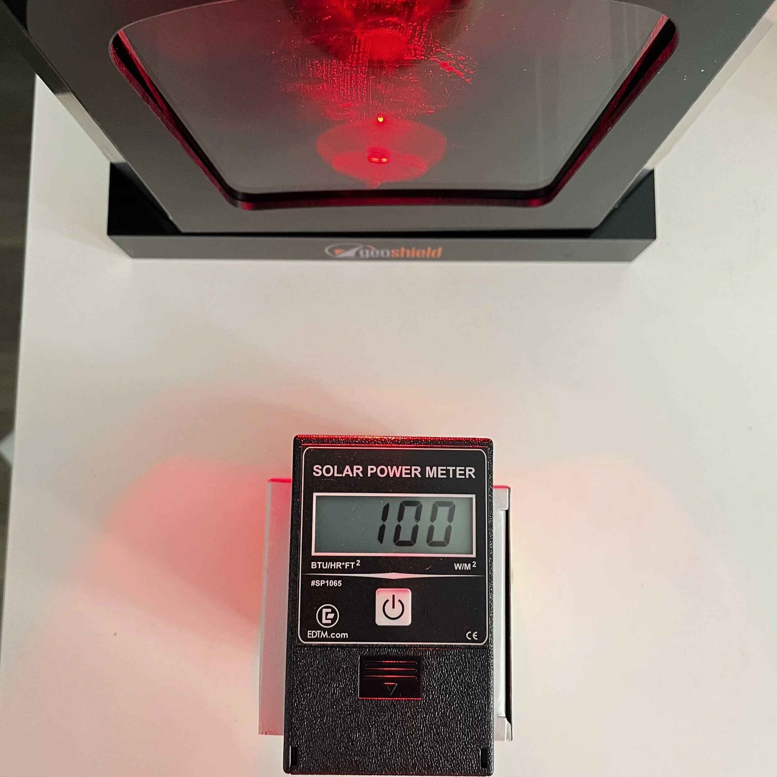 A solar power meter device on a white surface displaying 100 BTU/hr*FT squared on its screen, with a red laser light pointing downward.