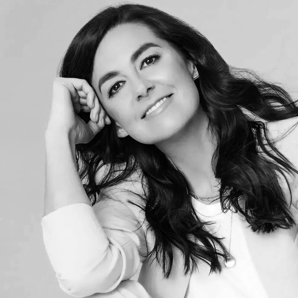 Black and white portrait of a woman with long dark hair, smiling, resting her head on her hand. Meredith Andrews.
